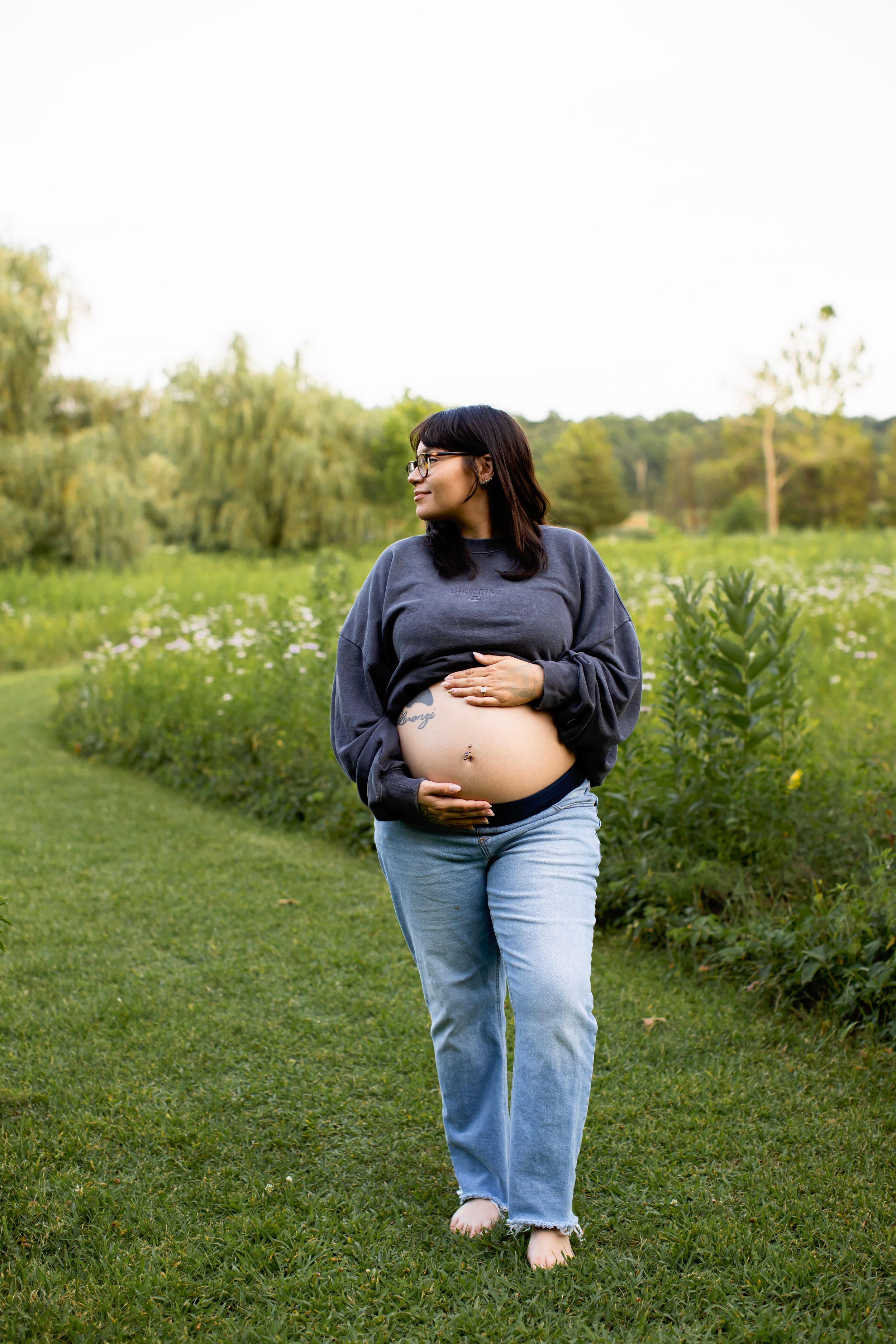 Expectant person in navy sweater and jeans cradles baby bump while standing in grassy field with willow trees.