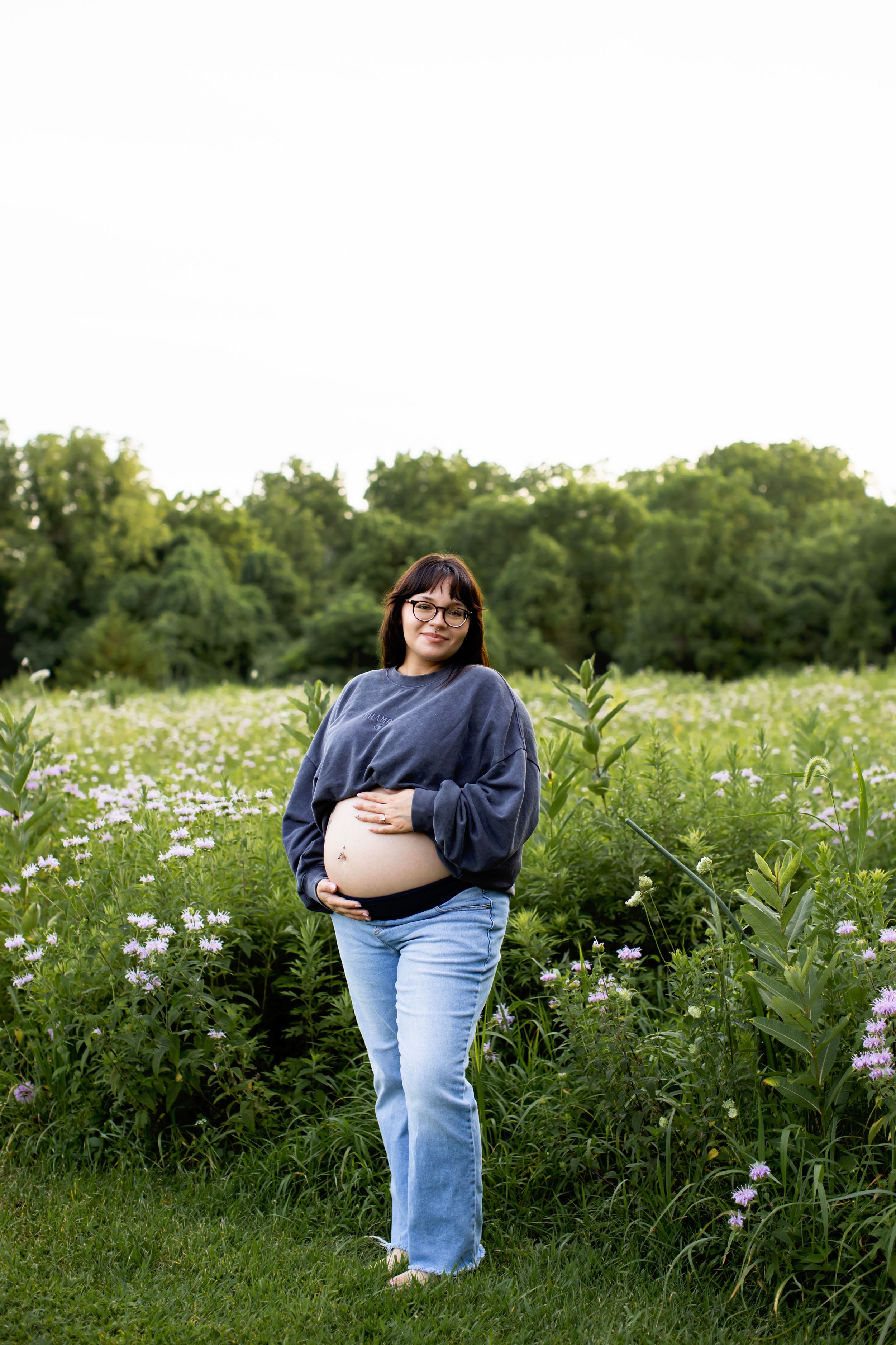 A pregnant woman in jeans and a navy sweater stands in a wildflower meadow during a maternity photoshoot.