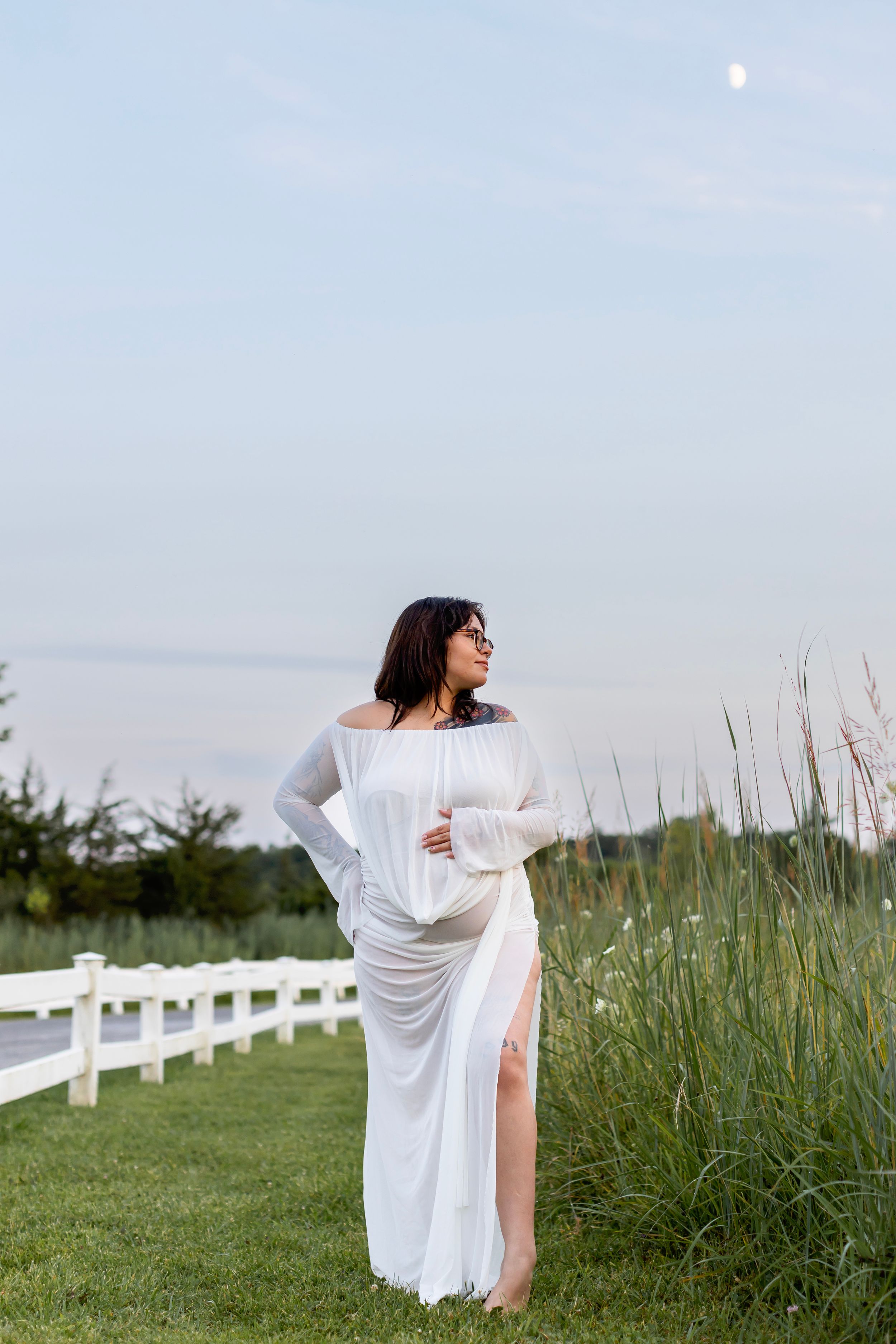 A person in a flowing white dress stands gracefully in a field with tall grass and white fence posts in the background.