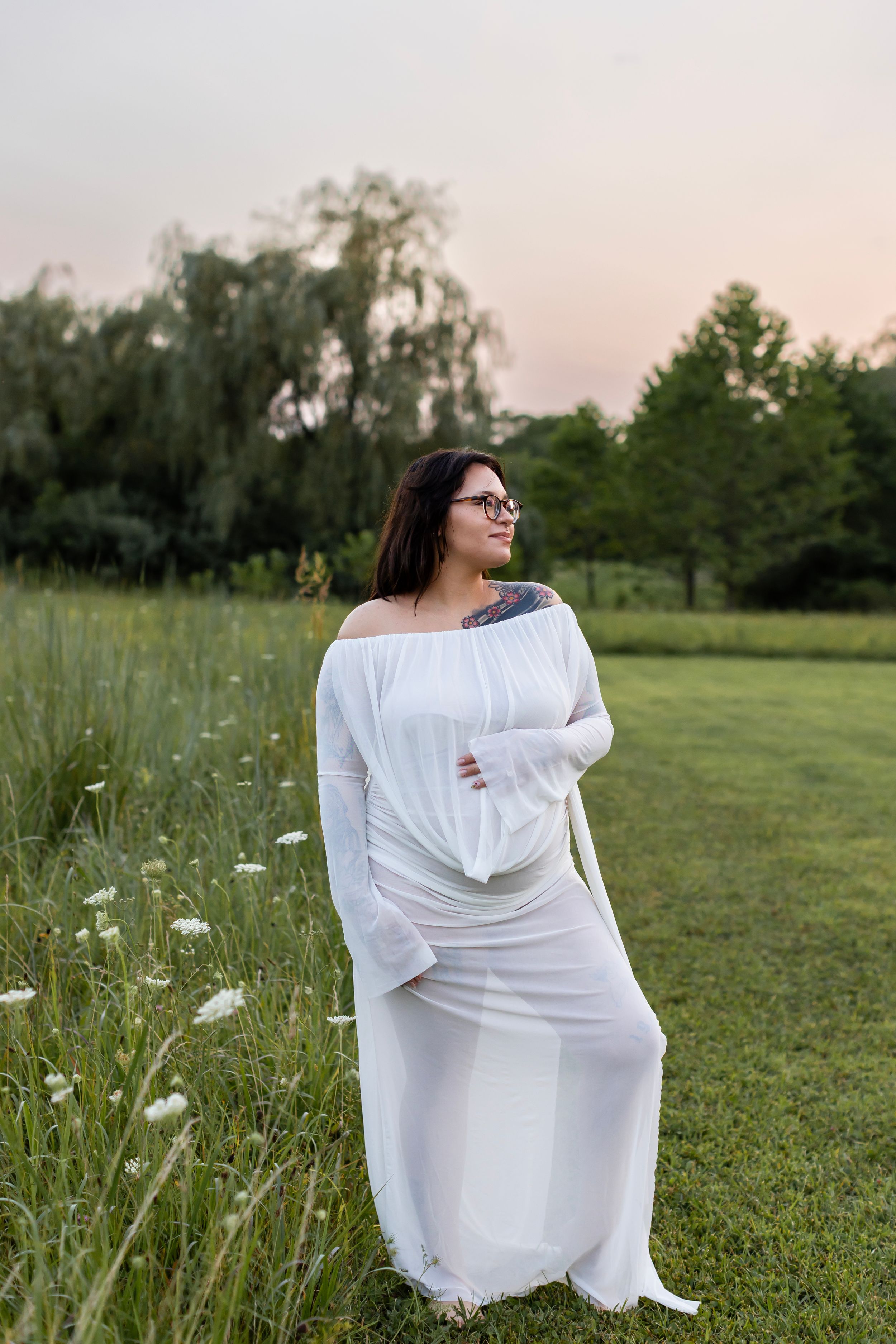 A person in a flowing white dress sits gracefully in a green meadow with wildflowers at sunset.