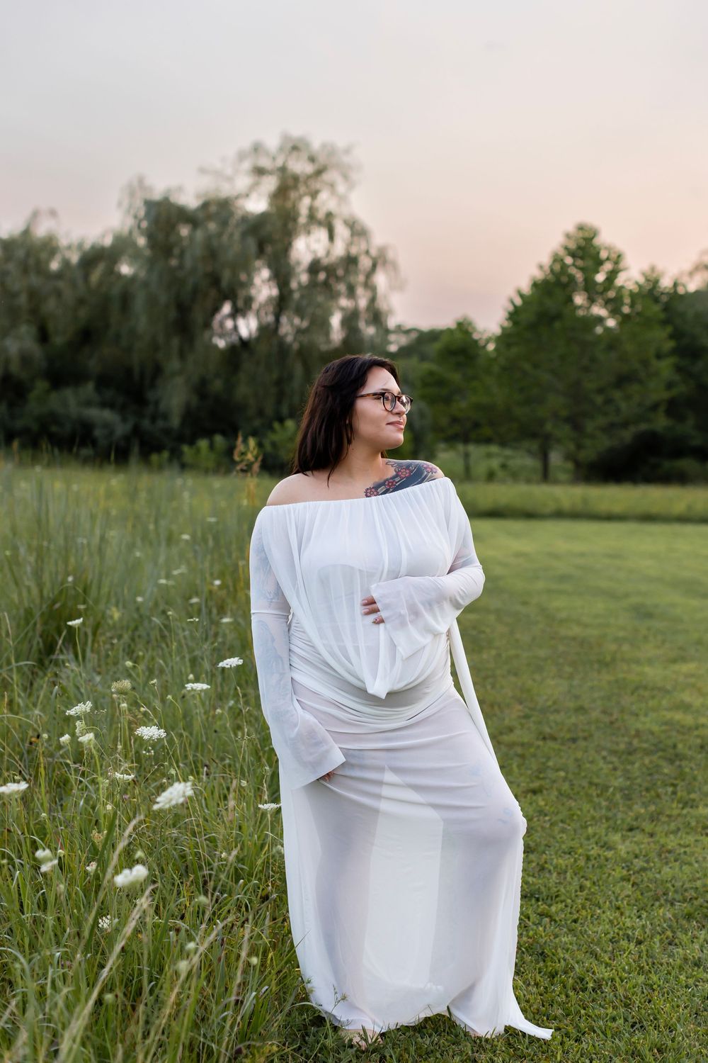 A person in a flowing white dress sits gracefully in a green meadow with wildflowers at sunset.