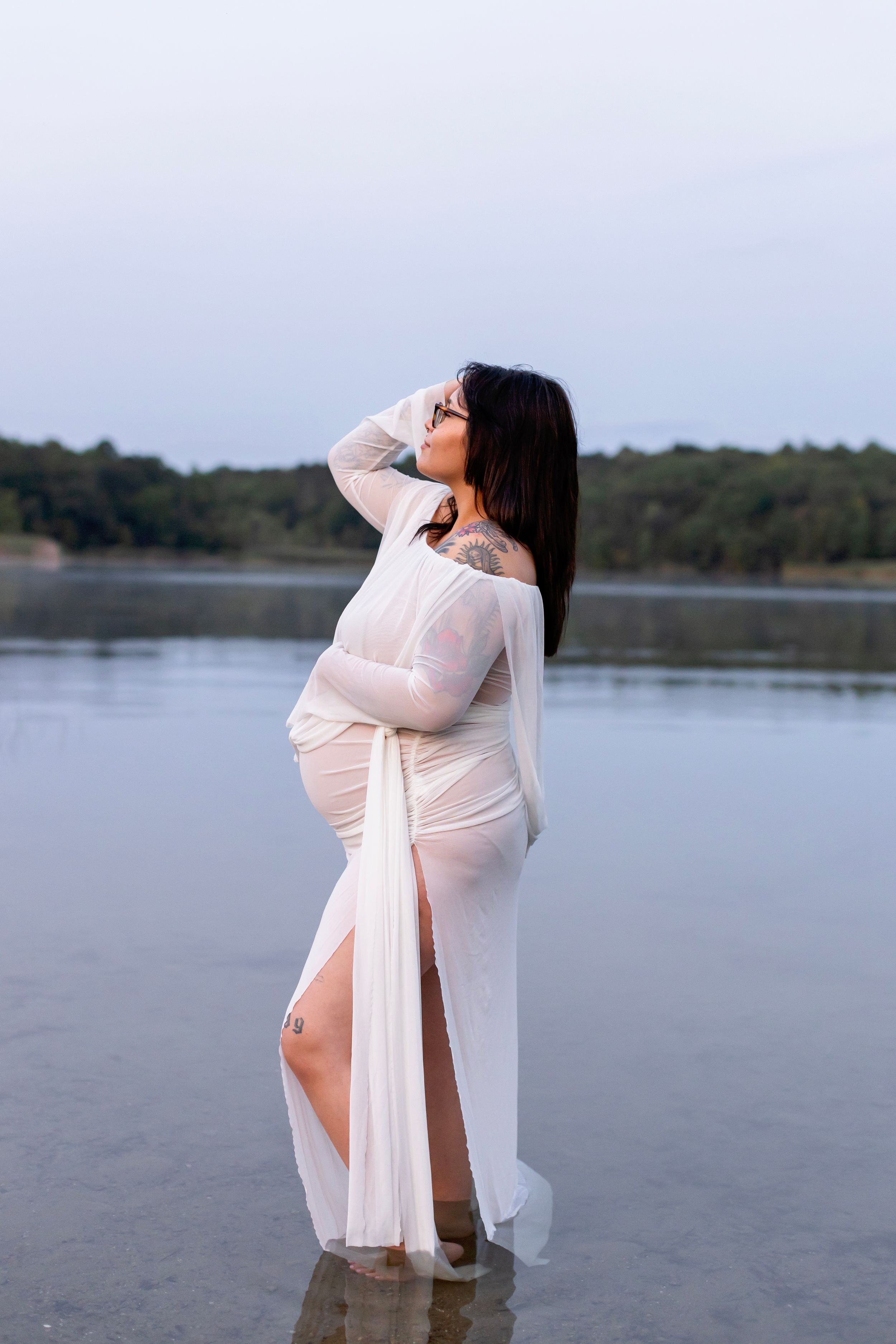 A maternity photoshoot by a serene lake features a figure in flowing white fabric against a tranquil water backdrop.