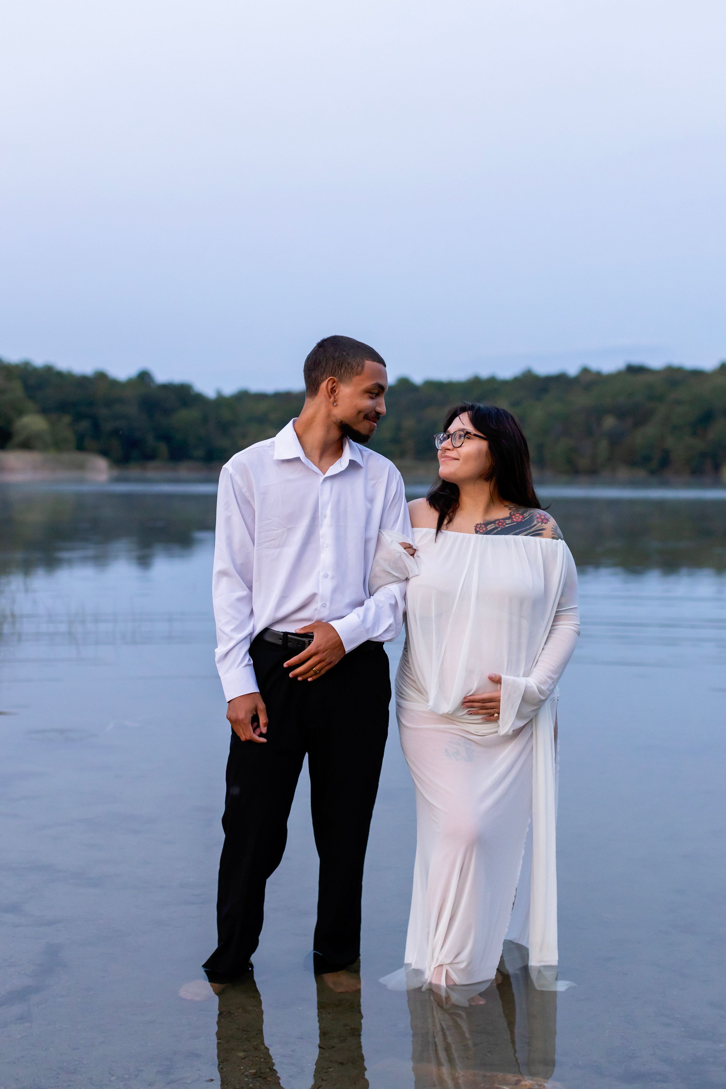 A couple in coordinating white and black outfits share a romantic moment by a serene lake during sunset.
