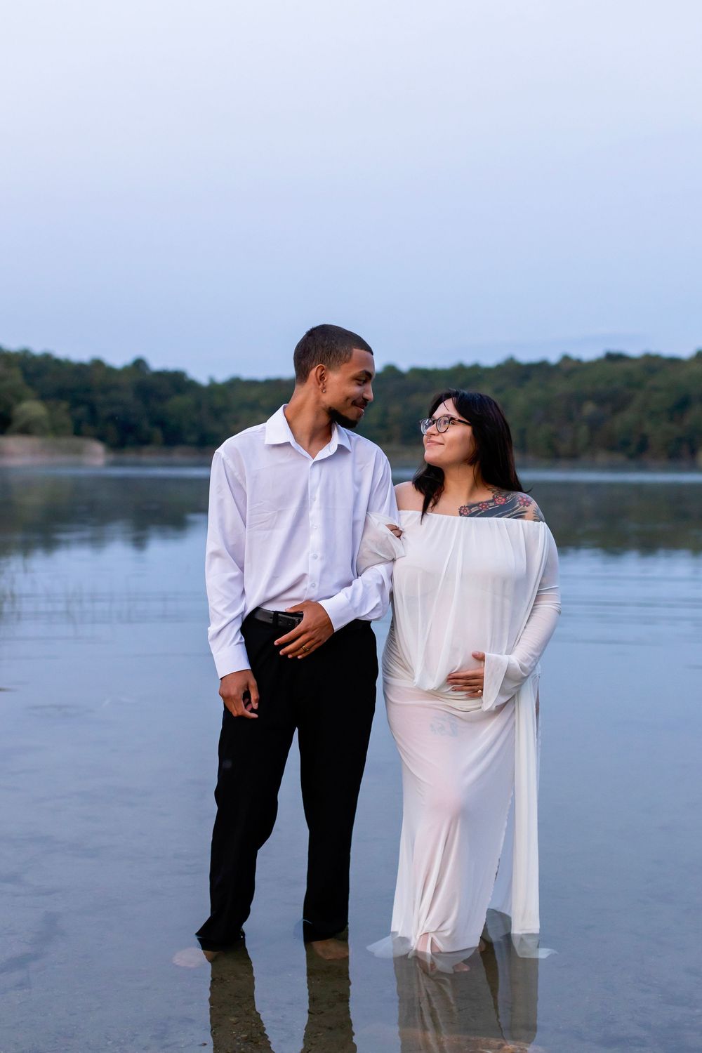 A couple in coordinating white and black outfits share a romantic moment by a serene lake during sunset.
