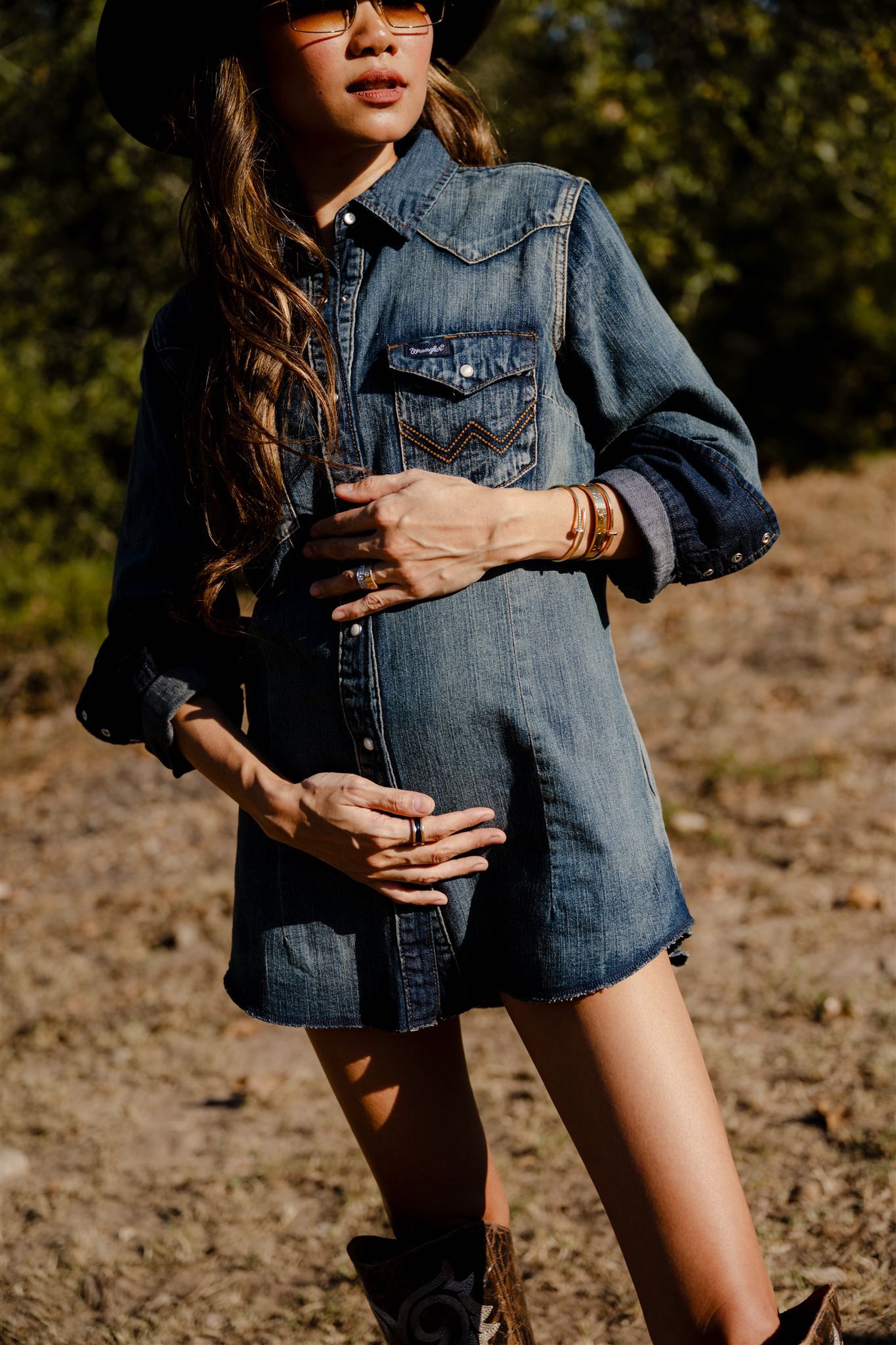 A woman wearing denim jacket and shorts poses in natural outdoor setting with sunlit rocky terrain in background.