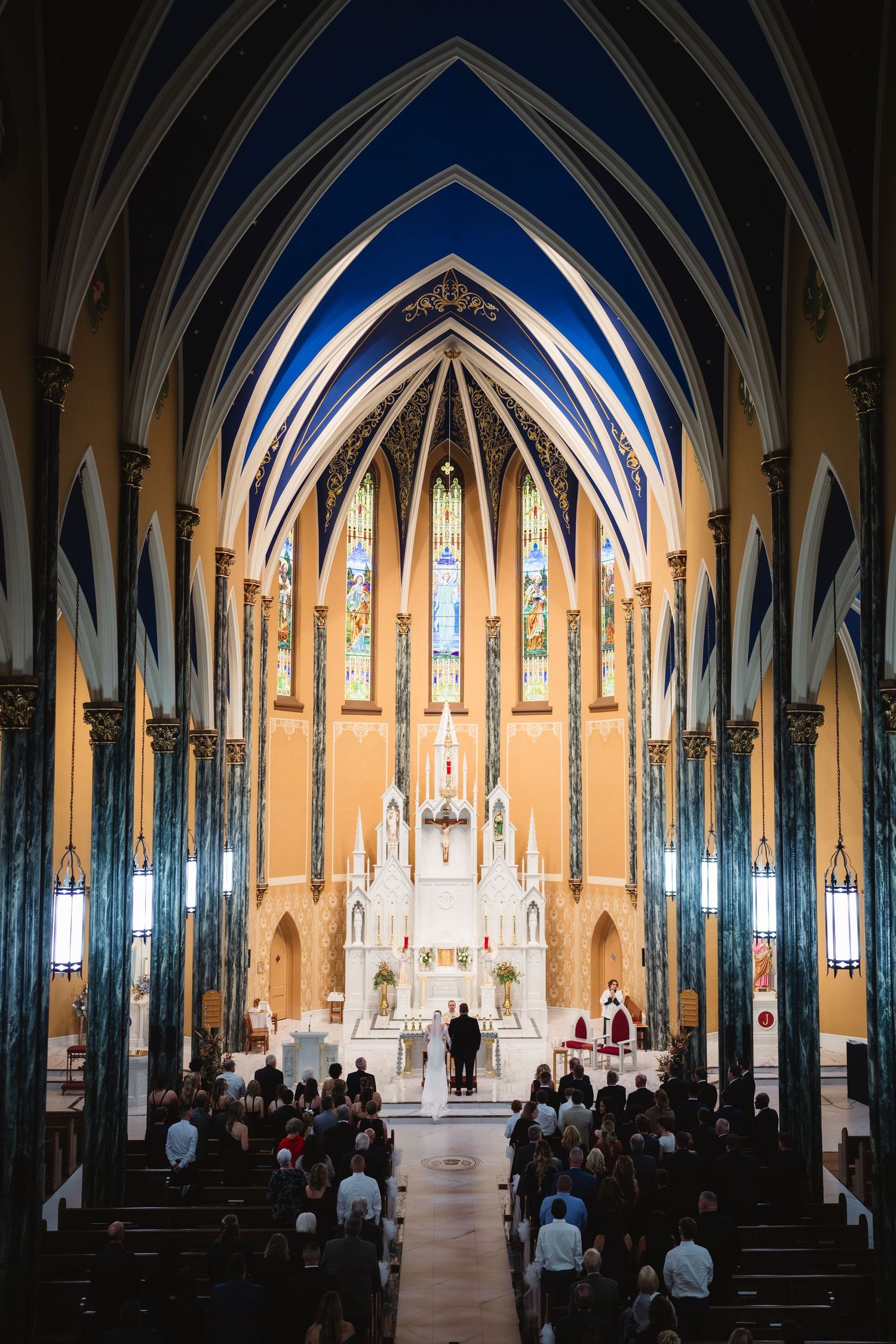 Interior view of a Gothic church with vaulted blue and gold ceiling, stained glass windows, and people attending service.