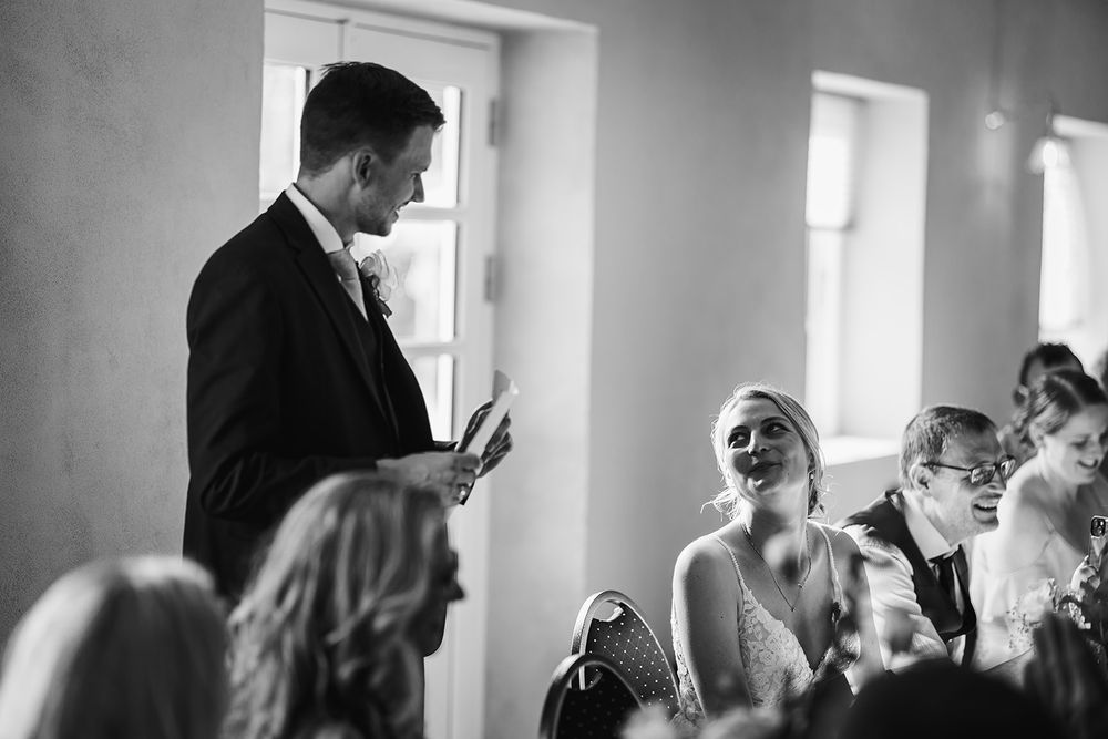 A black and white wedding ceremony scene showing guests seated in rows with natural light streaming through windows.