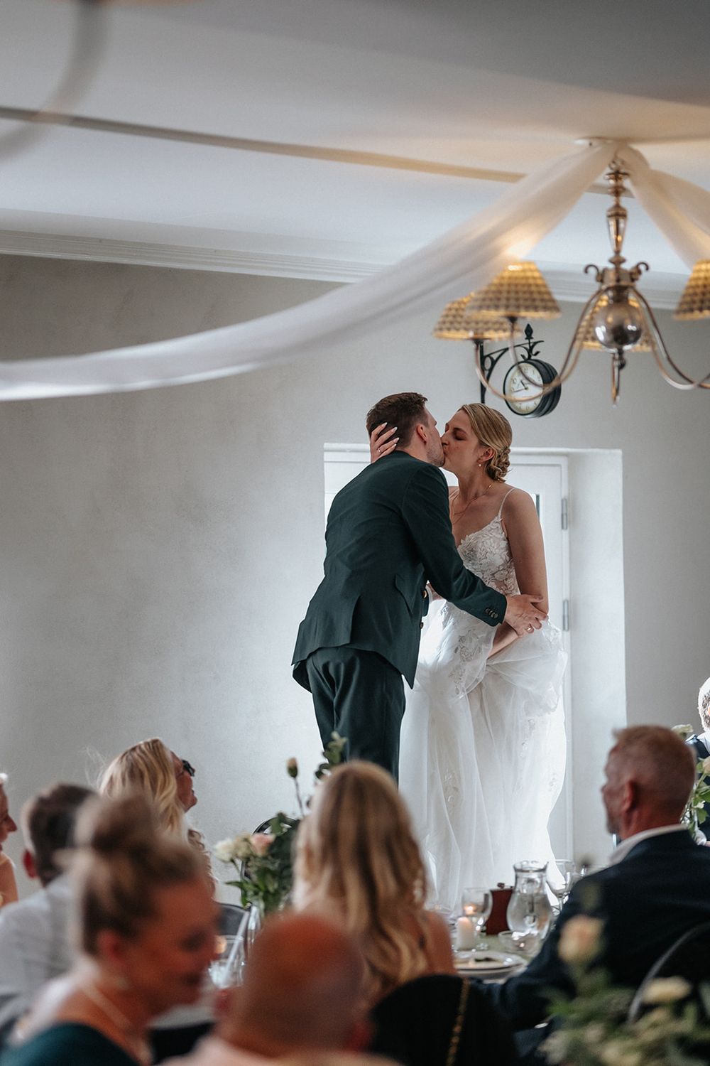 A romantic first kiss moment between newlyweds at a wedding reception with guests looking on.