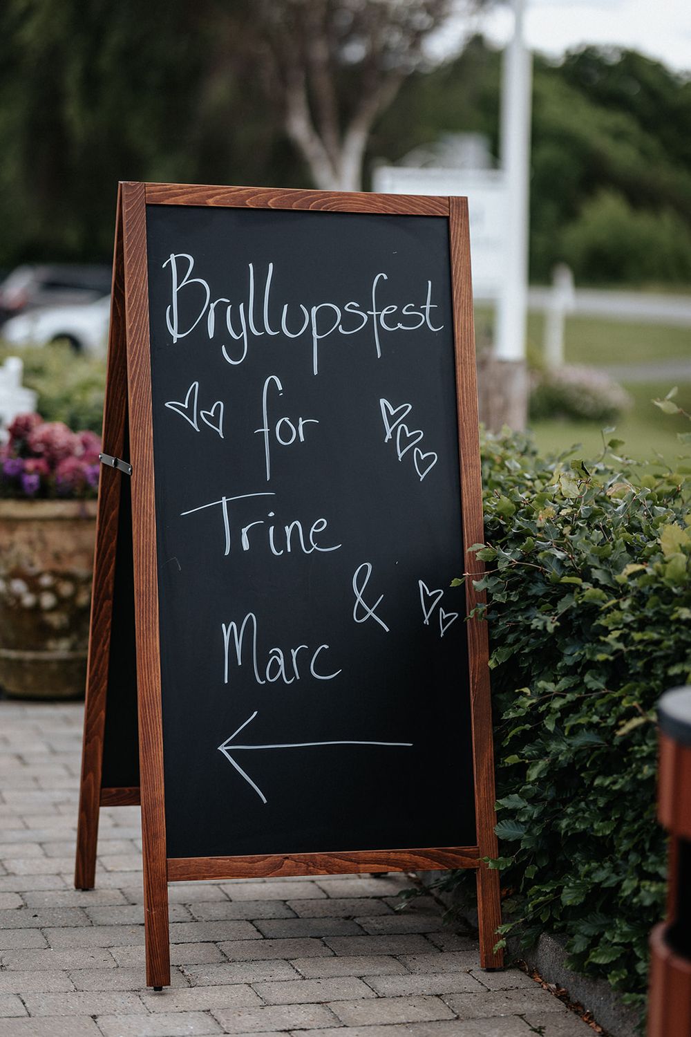 A wooden A-frame chalkboard sign displays 'Bryllupsfest' with hearts and an arrow on a brick pathway near greenery.