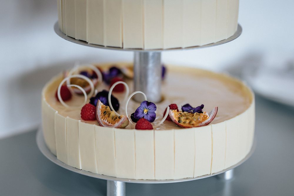 A white tiered cake stand displays an assortment of colorful macarons and desserts on a light surface.