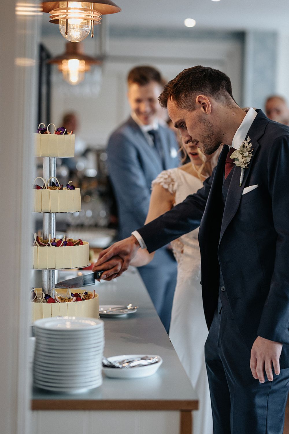 Wedding guests in formal attire gather around an elegant white tiered cake at an indoor reception venue.