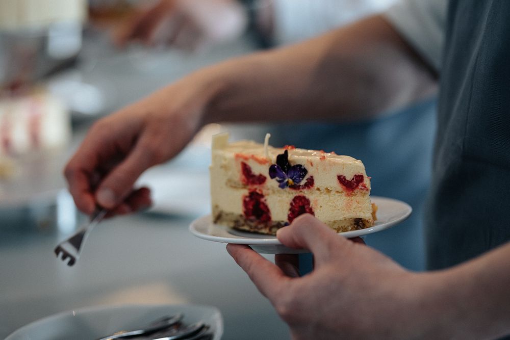 Several hands reaching for slices of cheesecake topped with berries on white plates at a gathering.