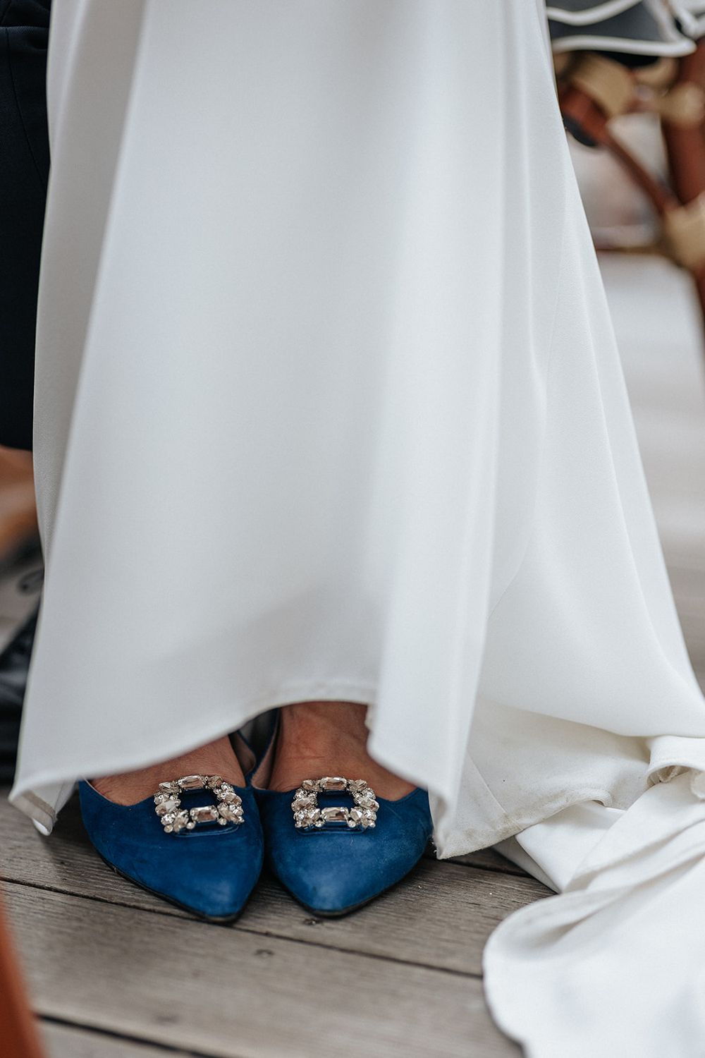 Blue suede wedding shoes with crystal embellishments peek out from under a flowing white bridal gown.
