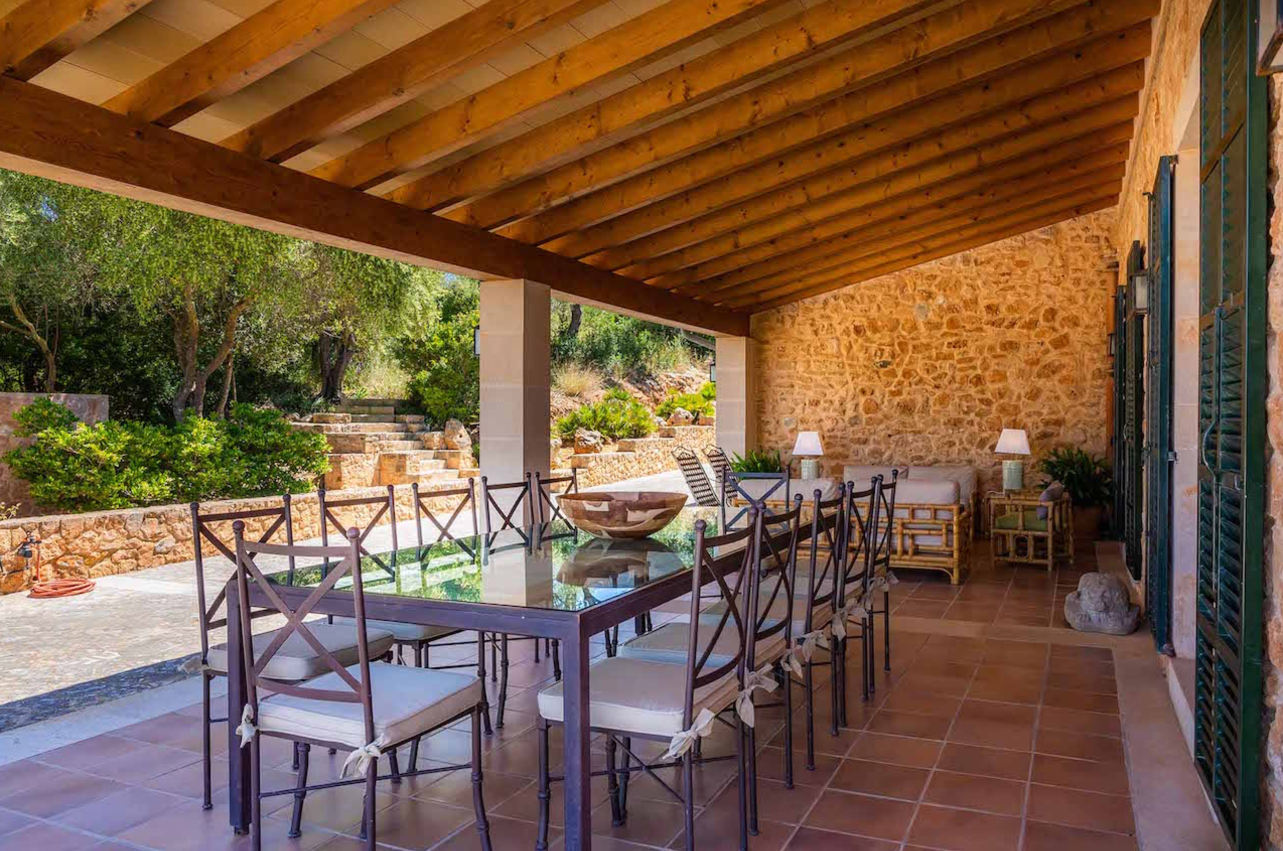 A rustic covered patio with wooden beams and stone walls features long dining tables set for outdoor dining.