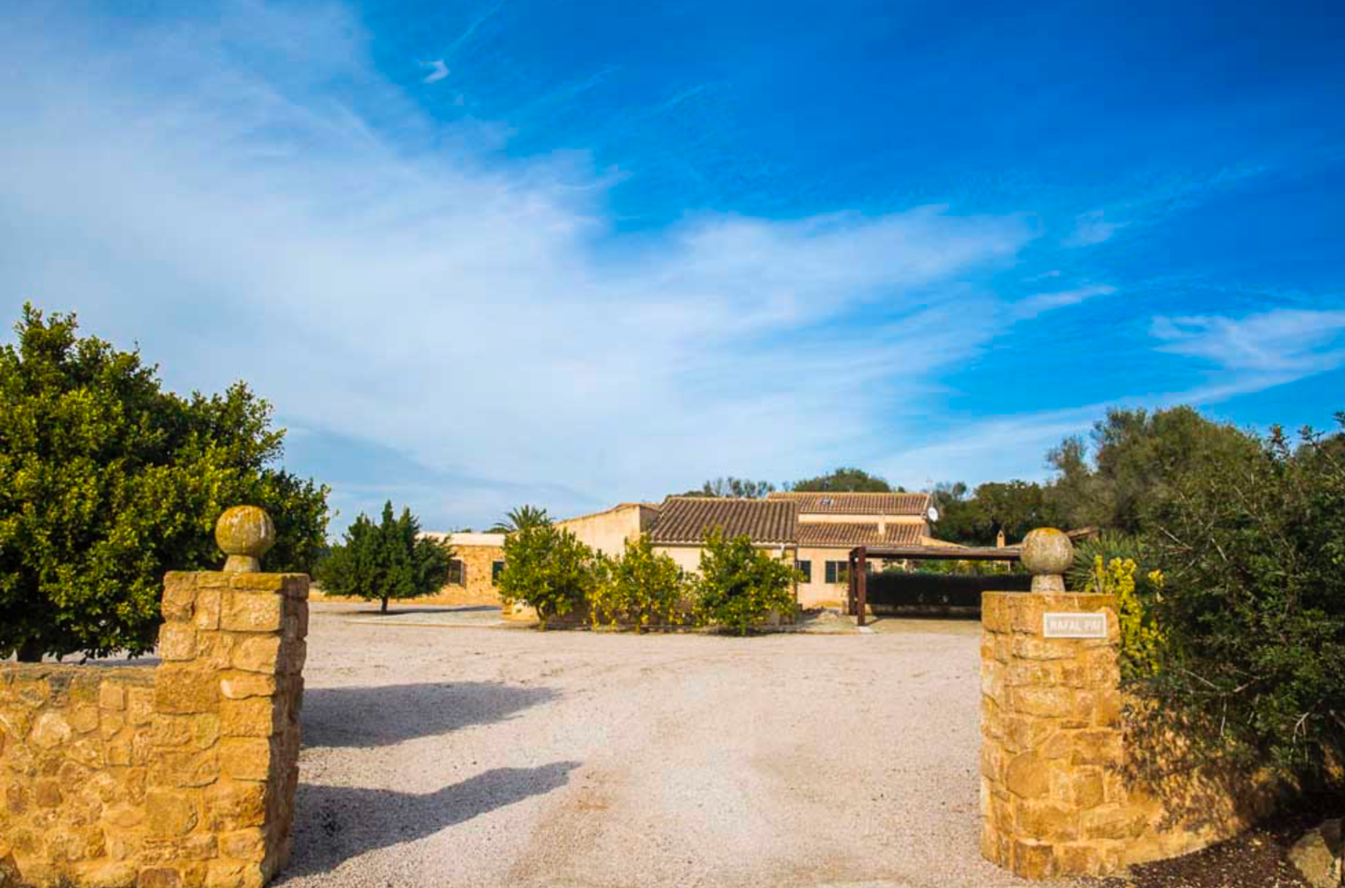 Stone entrance pillars and winding driveway lead to a Mediterranean style estate under bright blue skies.