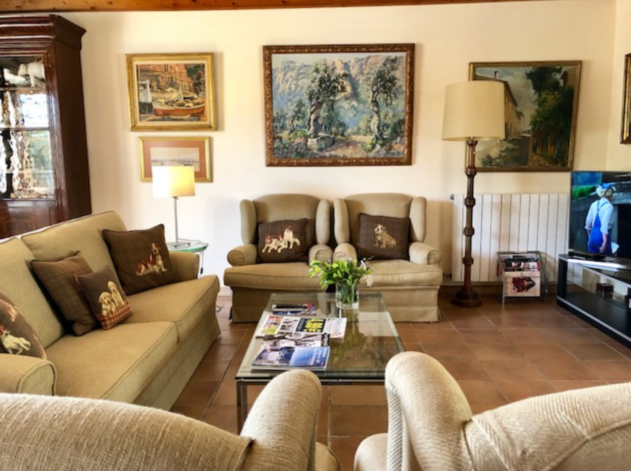 A panoramic view of a cozy living room with beige furniture, artwork on walls, and warm lighting throughout.