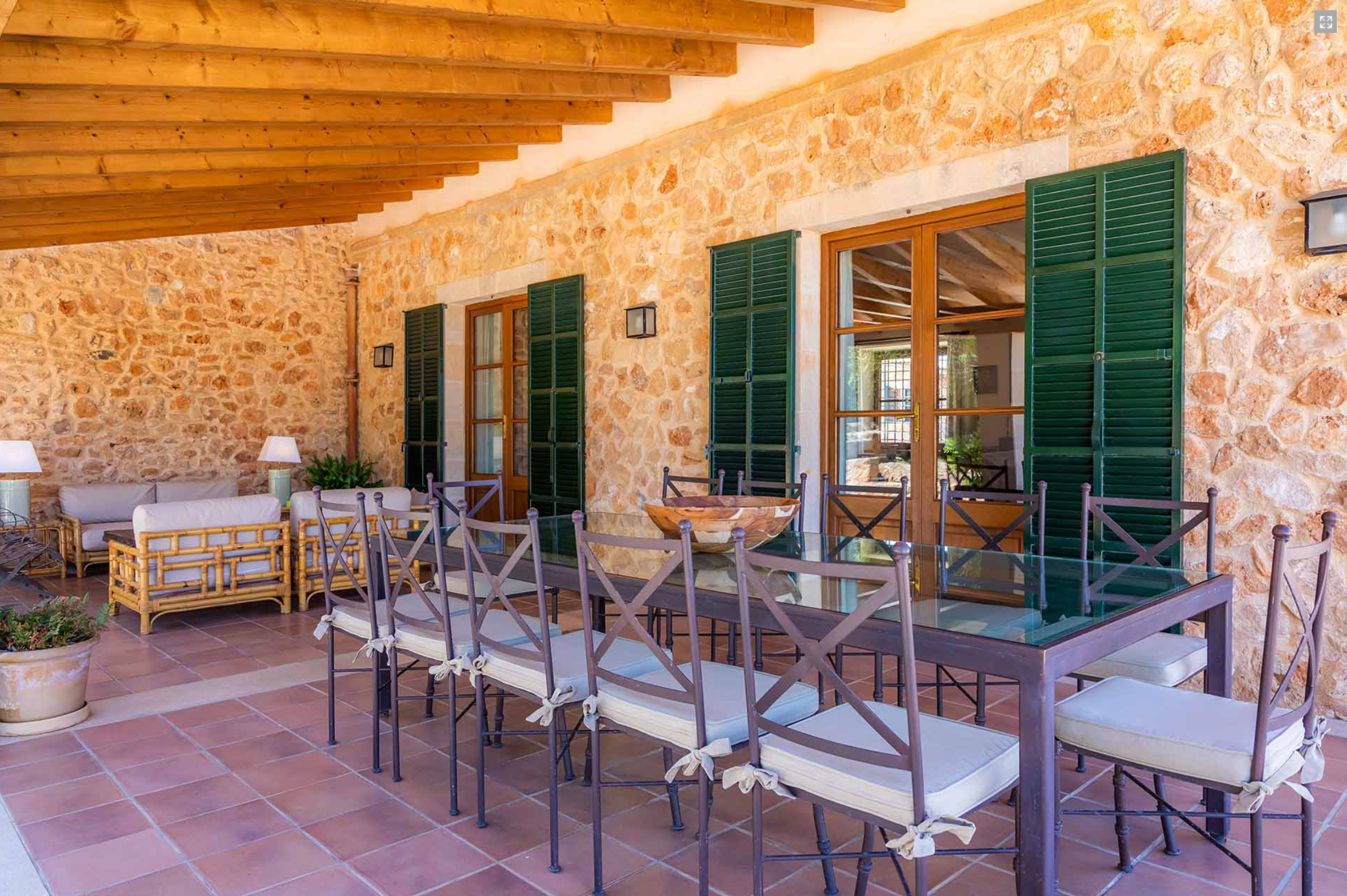 Stone patio with white cushioned seating and green shuttered windows along Mediterranean style wall.