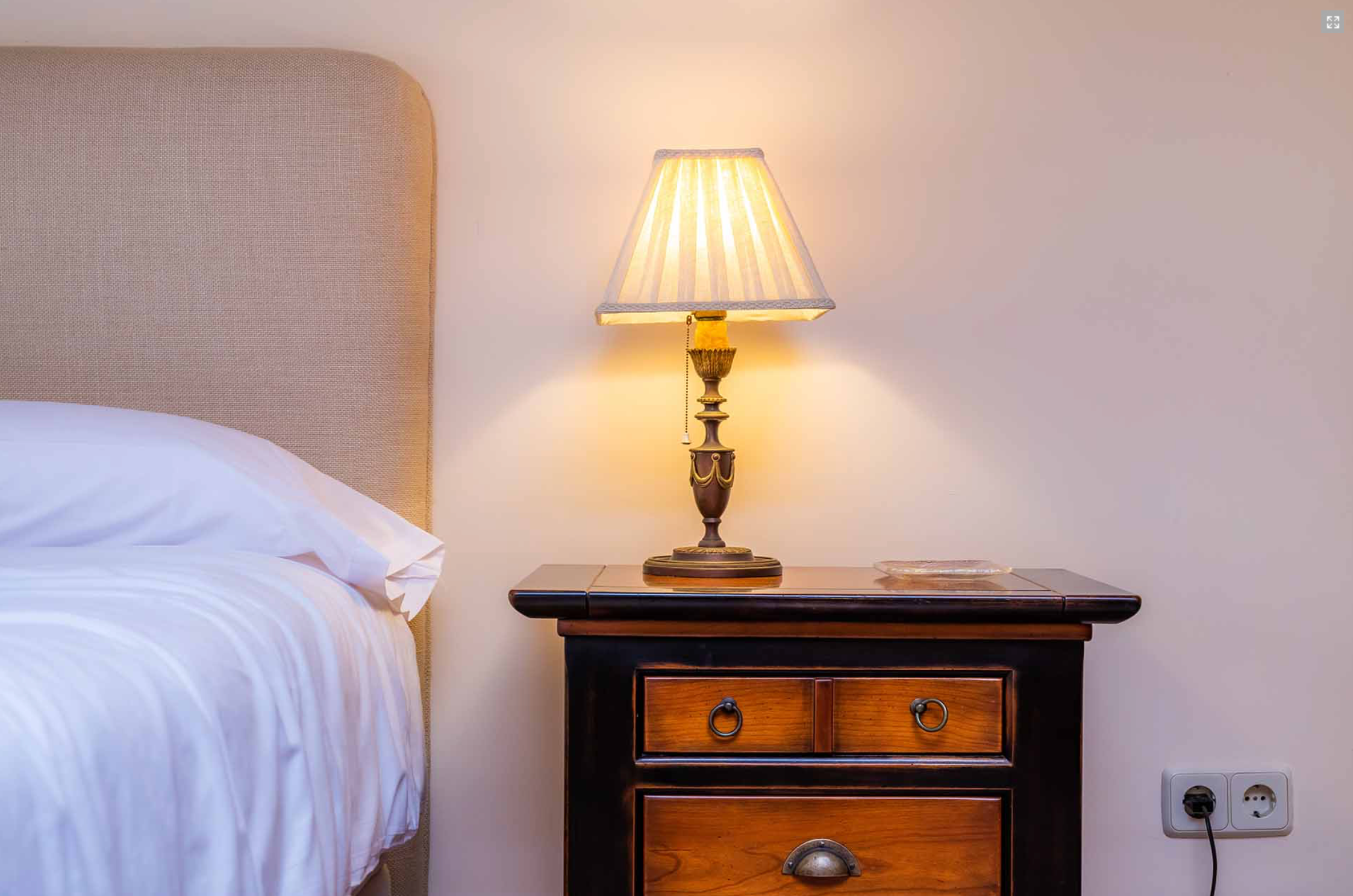 Cozy bedroom corner with antique nightstand, traditional table lamp and white bedding in warm lighting.