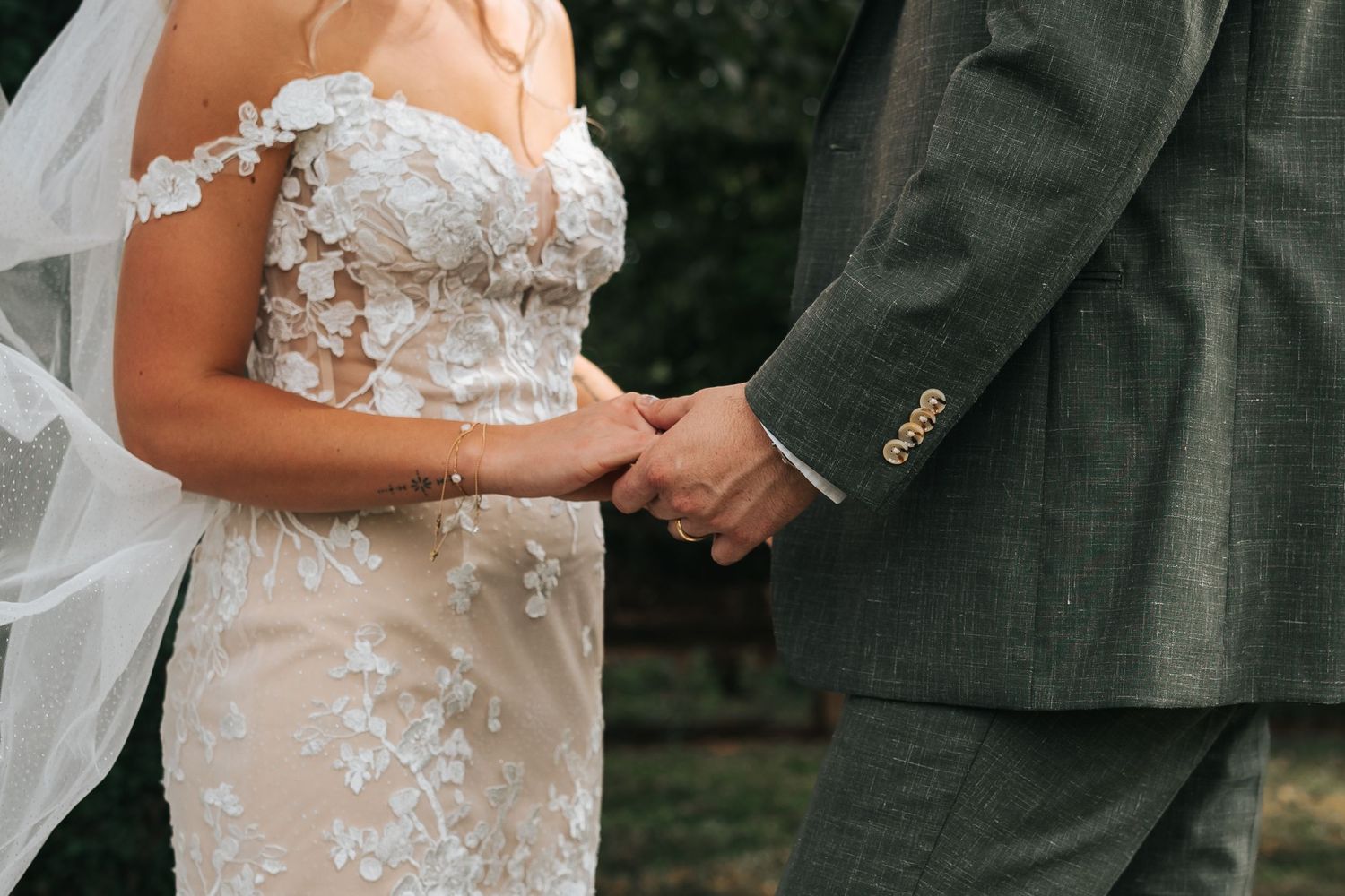 Couple in wedding attire holding hands during outdoor ceremony with white floral lace dress details.