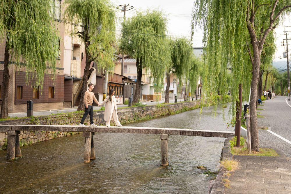 Couple crossing a small stone bridge over a canal lined with weeping willow trees in Kyoto, Japan.