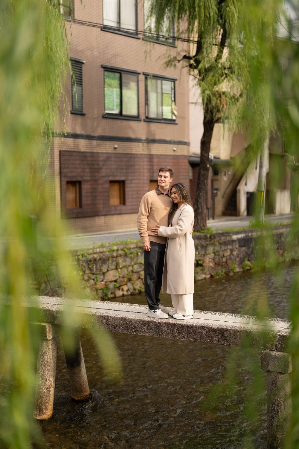Couple standing together on a canal walkway framed by weeping willow trees in Kyoto, Japan.