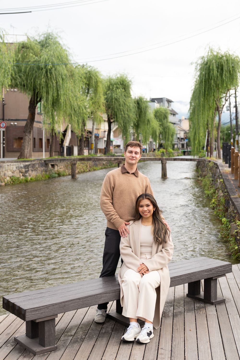 Couple posing on a wooden deck bench beside a tranquil canal with weeping willows in Kyoto, Japan.