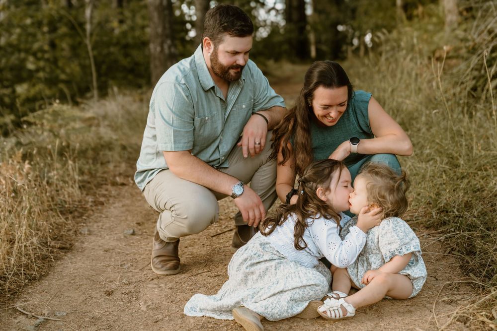 Family of four shares tender moment on forest trail, parents watching two young daughters in floral dresses.