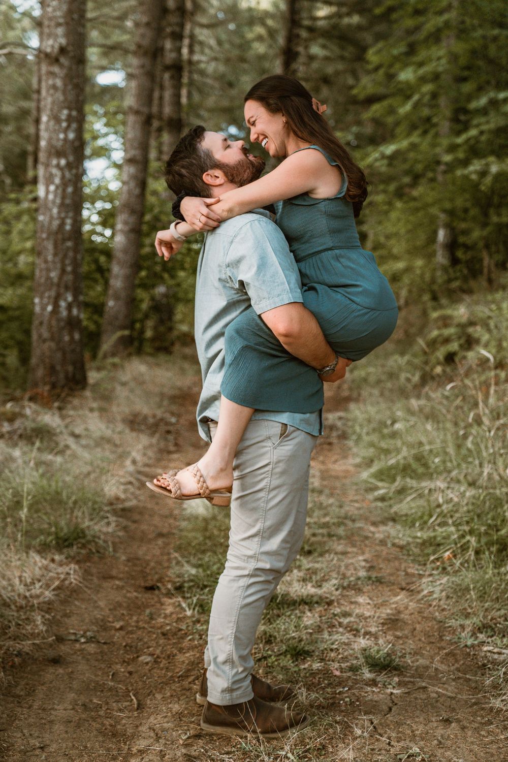 Bearded man lifts smiling woman in teal dress in a romantic embrace on a forest trail.