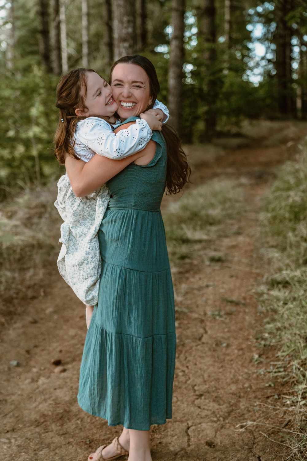 Joyful mother in teal dress holds daughter who gives her a kiss on the cheek on a forest path.
