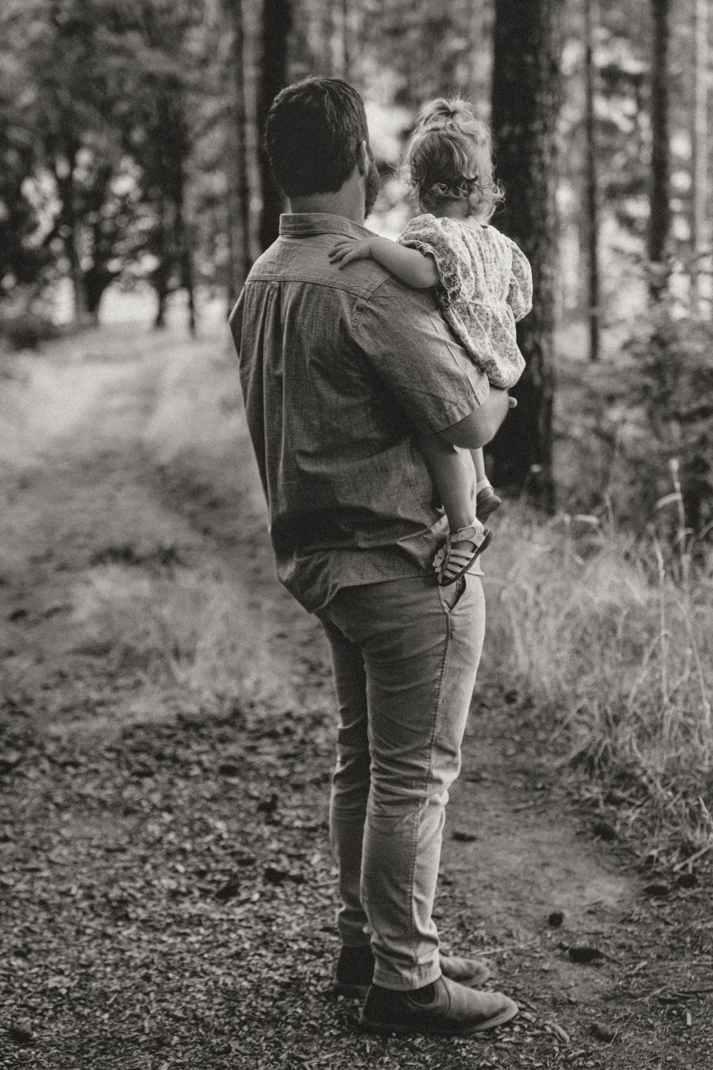 Black and white photo of father carrying young child on a peaceful wooded forest trail.