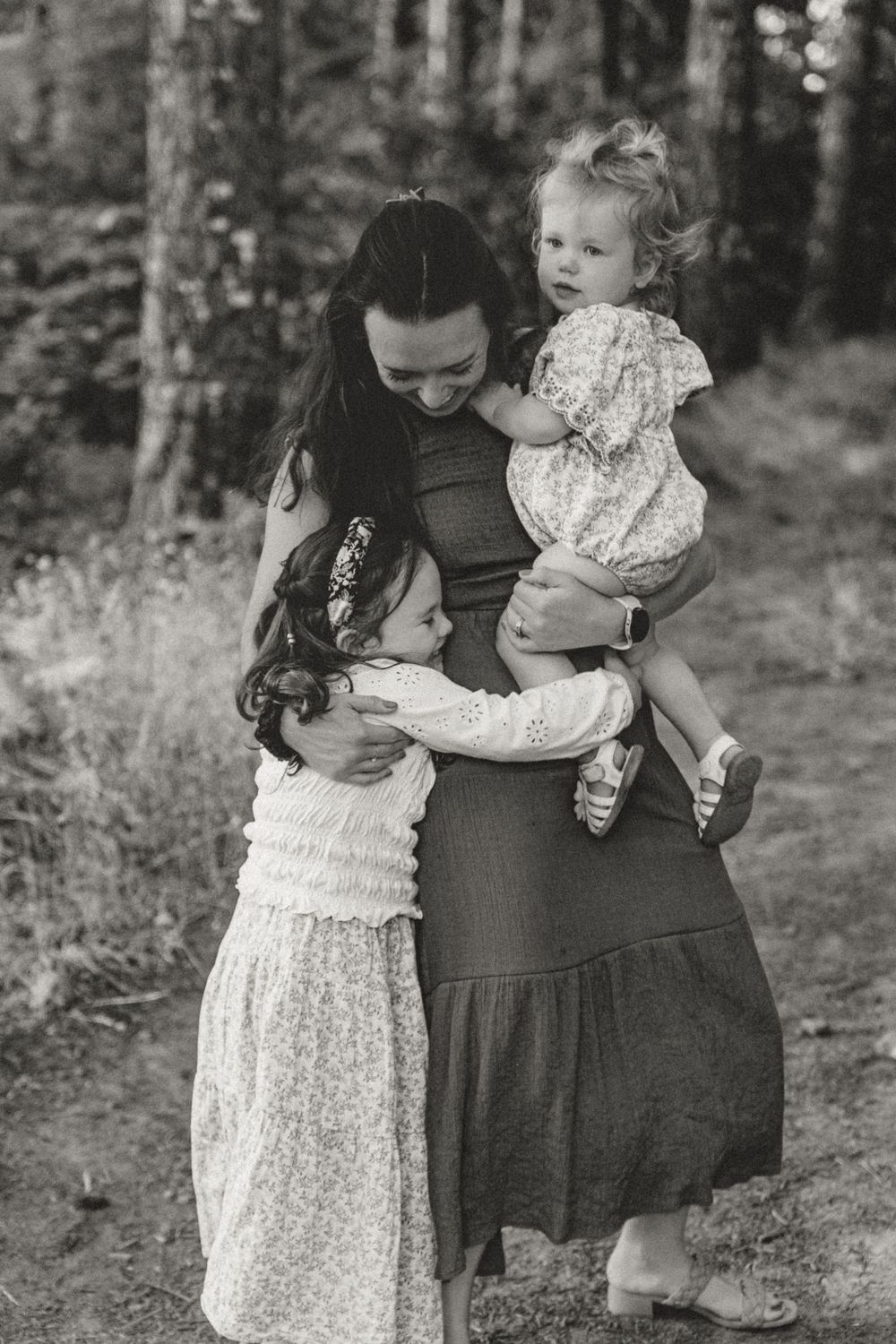 Black and white portrait of mother embracing two young daughters in a tender forest setting.