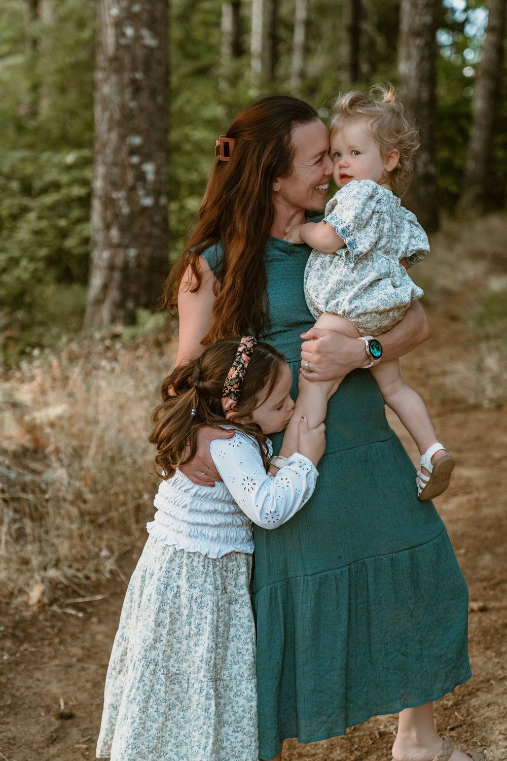 Redheaded mother in teal dress holds toddler while older daughter hugs her in a pine forest.
