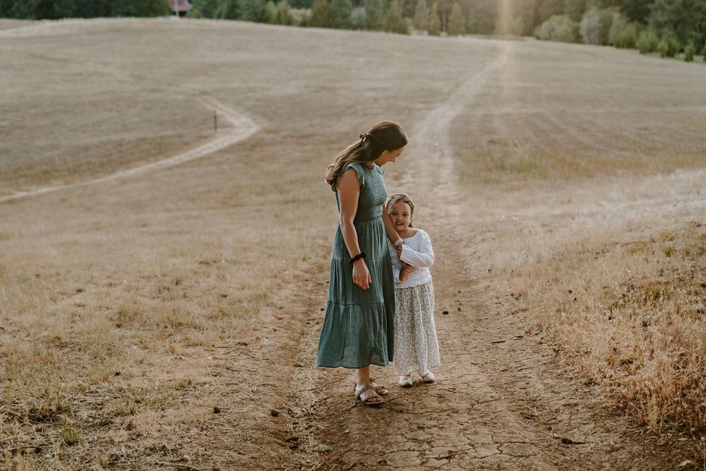 A mother in a teal dress walks with her young daughter in a white dress on a sunny dirt path in a field.