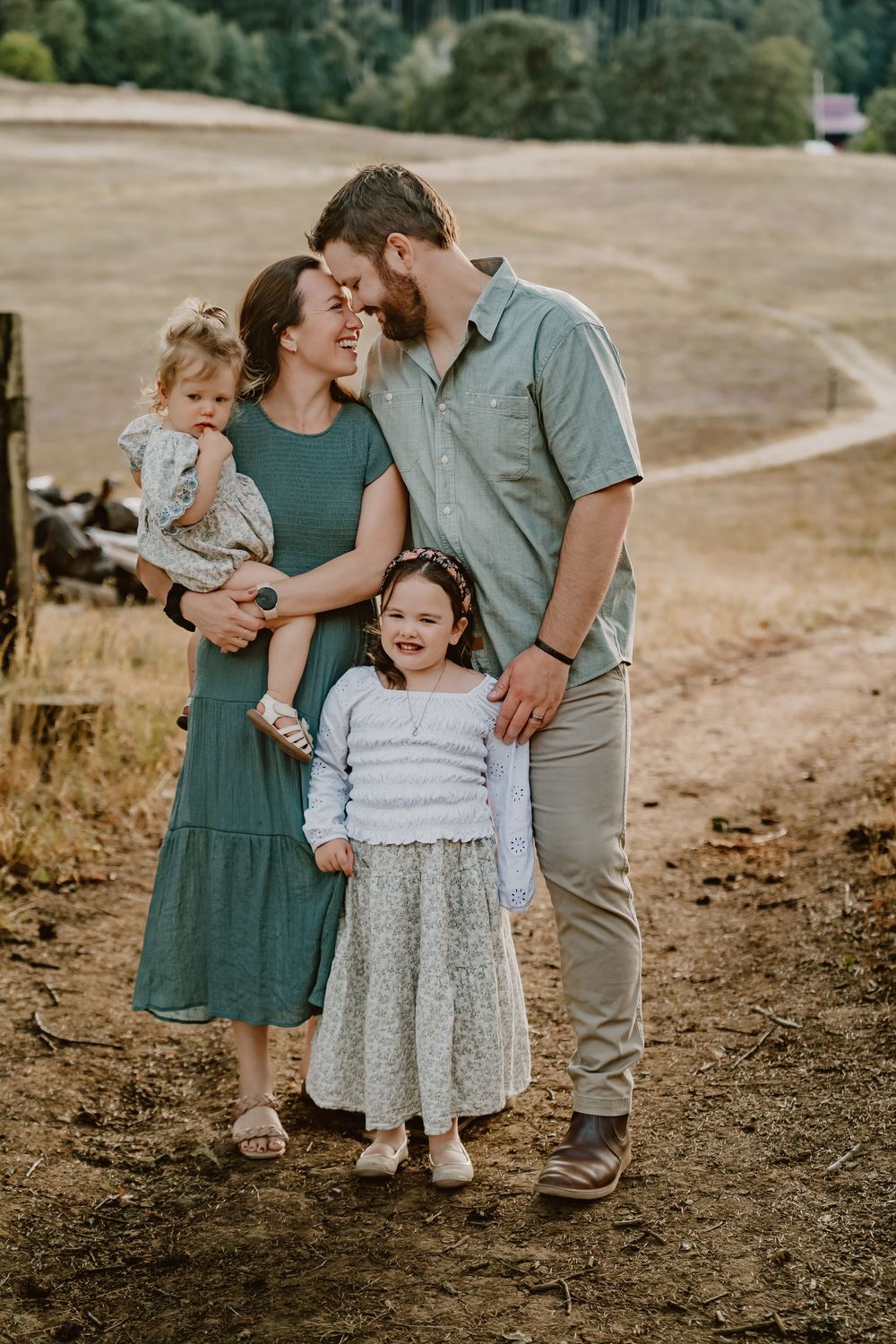 A happy family of four poses on a rural dirt path, parents sharing a kiss while holding their two young daughters.
