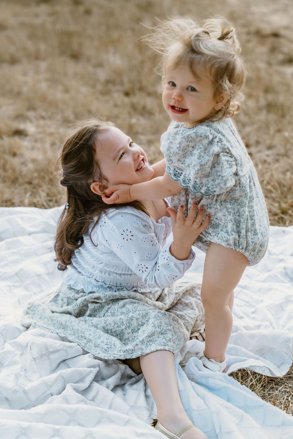 Two sisters laugh and play together on a light blanket in a golden field, sharing a joyful moment.