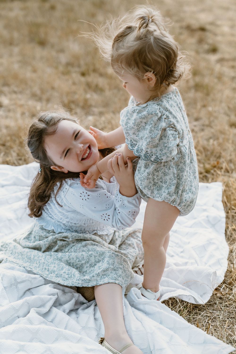 Two young sisters share a playful moment on a blanket in a field, the older girl laughing with delight.