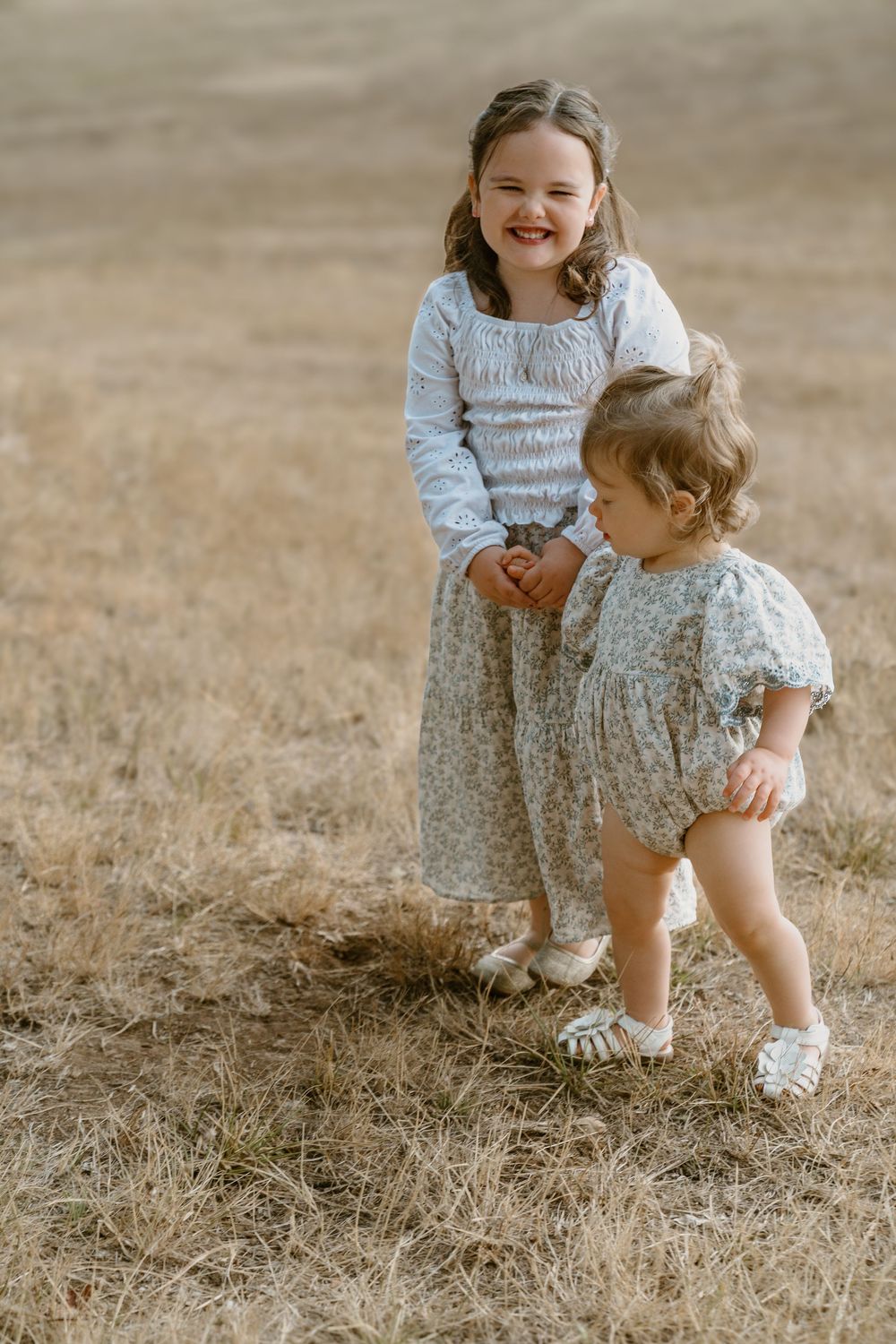 A smiling older sister in a white dress stands with her toddler sister in a floral romper in a dry field.
