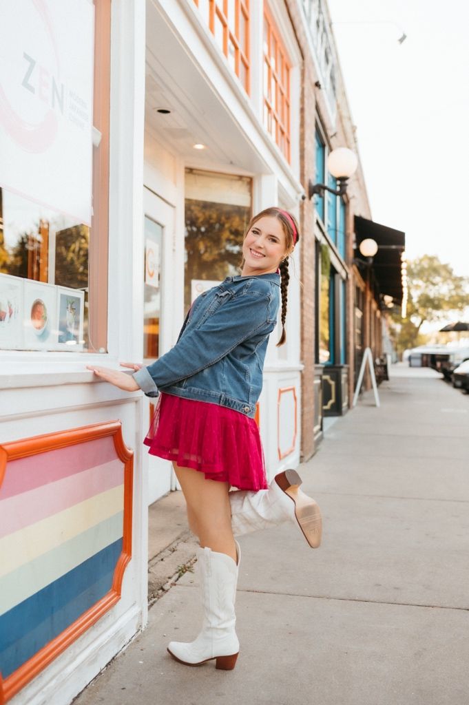 Person wearing denim and red skirt poses playfully near white storefront.