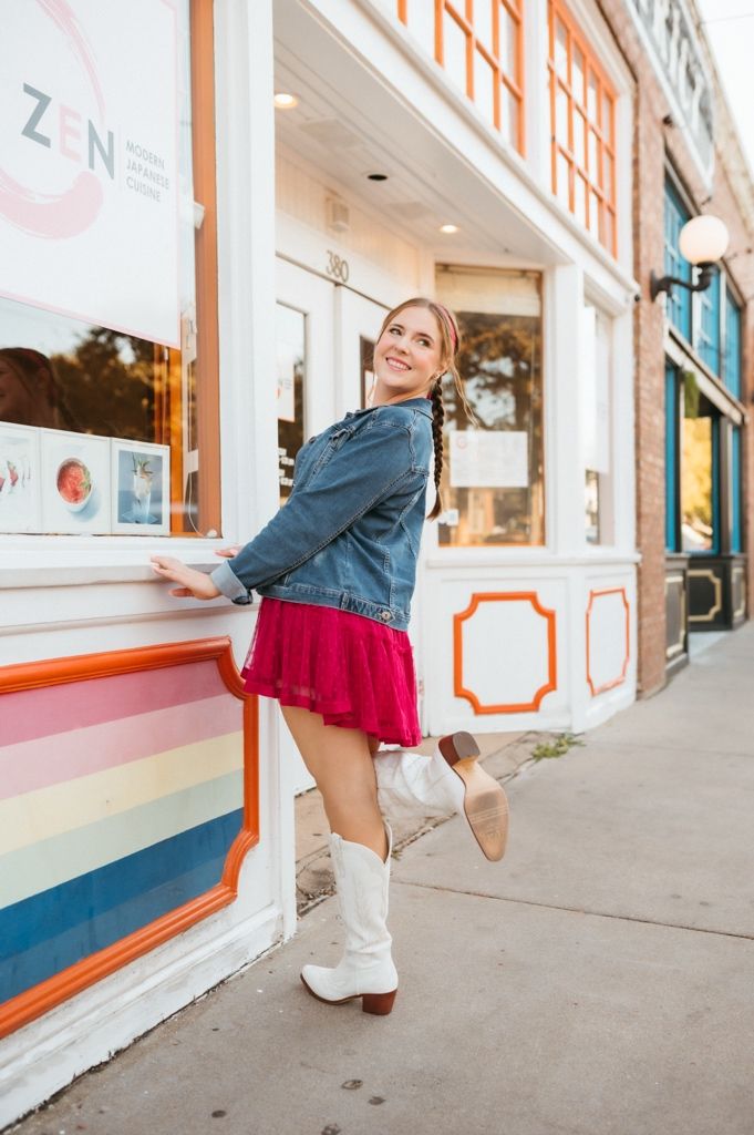 Playful street fashion photo with white boots, red dress and denim jacket against colorful storefront.