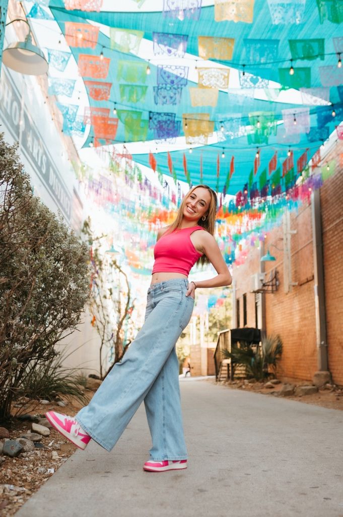 Casual fashion pose under colorful papel picado banners in an alley with pink sneakers.