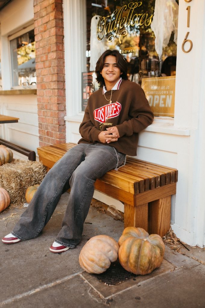 A person in a brown hoodie sits on a wooden bench next to autumn pumpkins outside a brick storefront.
