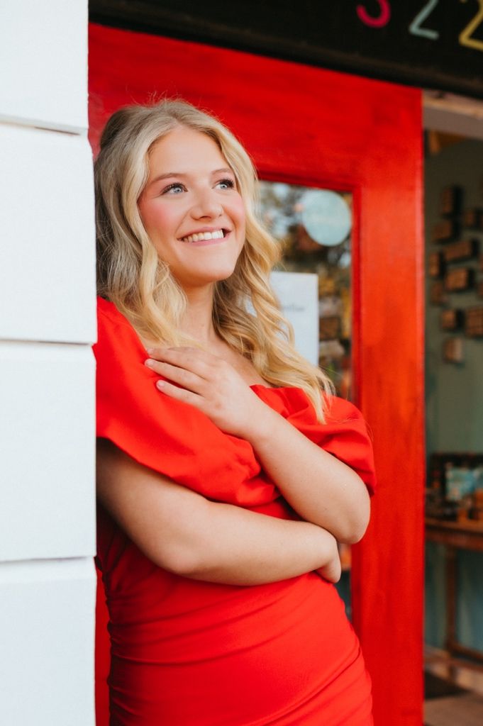 Portrait against a red storefront wearing a bright red dress with a joyful expression.