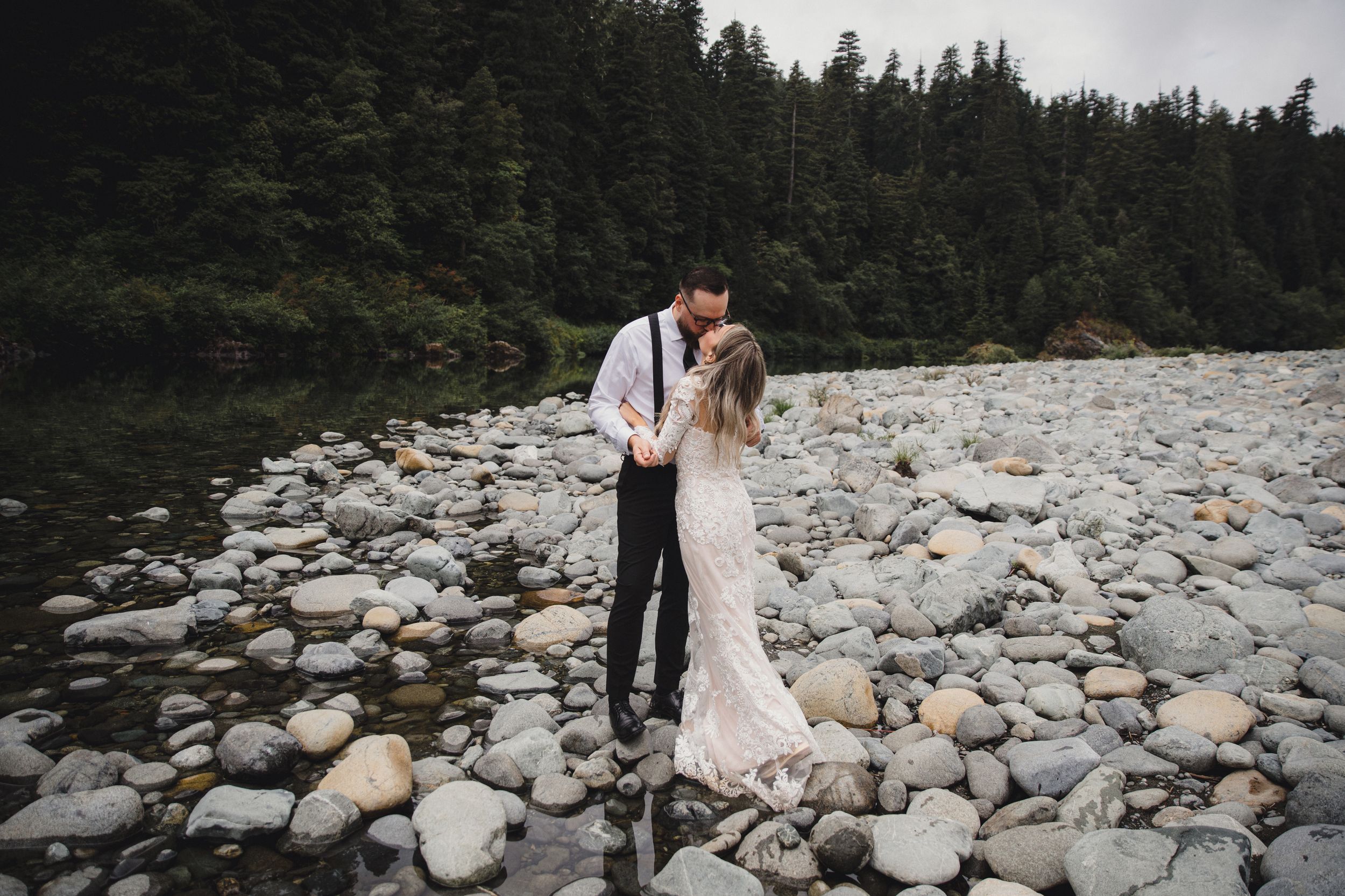 A couple shares romantic moments on a rocky beach surrounded by evergreen forest in the Pacific Northwest.
