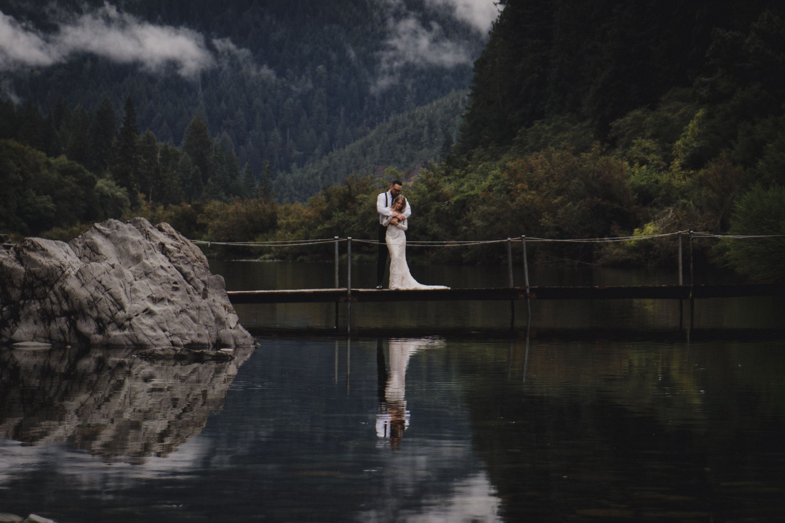 A couple stands on a wooden bridge over a tranquil mountain lake with dramatic reflections and misty peaks in the background.