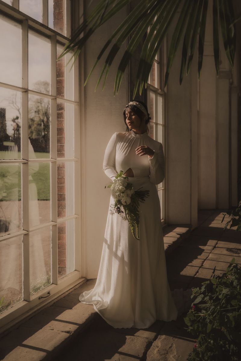 A bride in a flowing white wedding dress stands by large windows in a sunlit room with indoor plants.