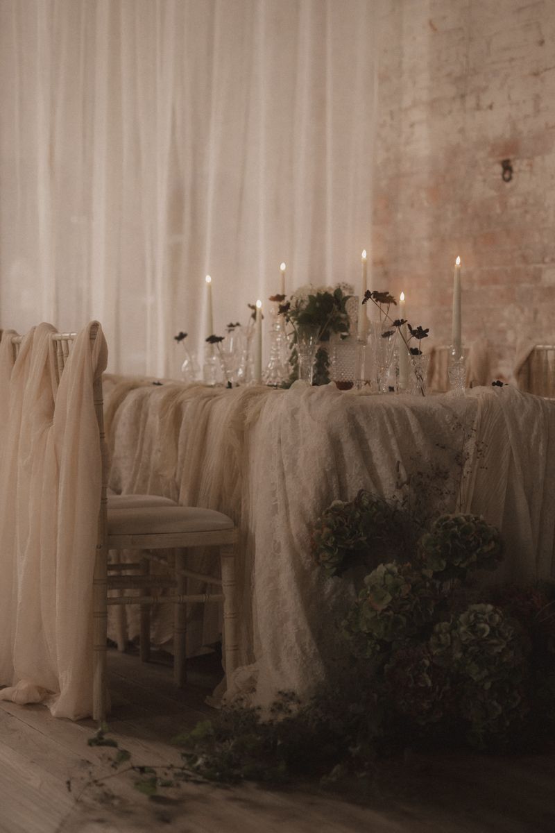 Elegant wedding table setting with white linen tablecloth, taper candles and greenery in a rustic venue with brick walls.