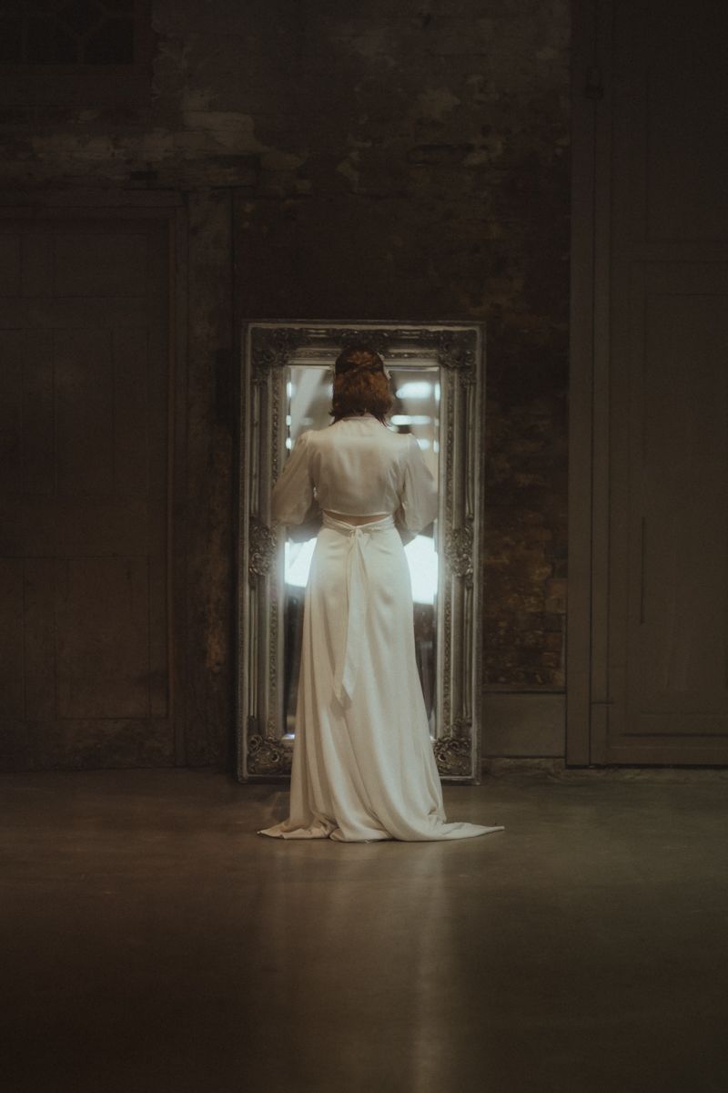 A figure in a white wedding dress sits in front of a bright window in a dark, moody industrial space.