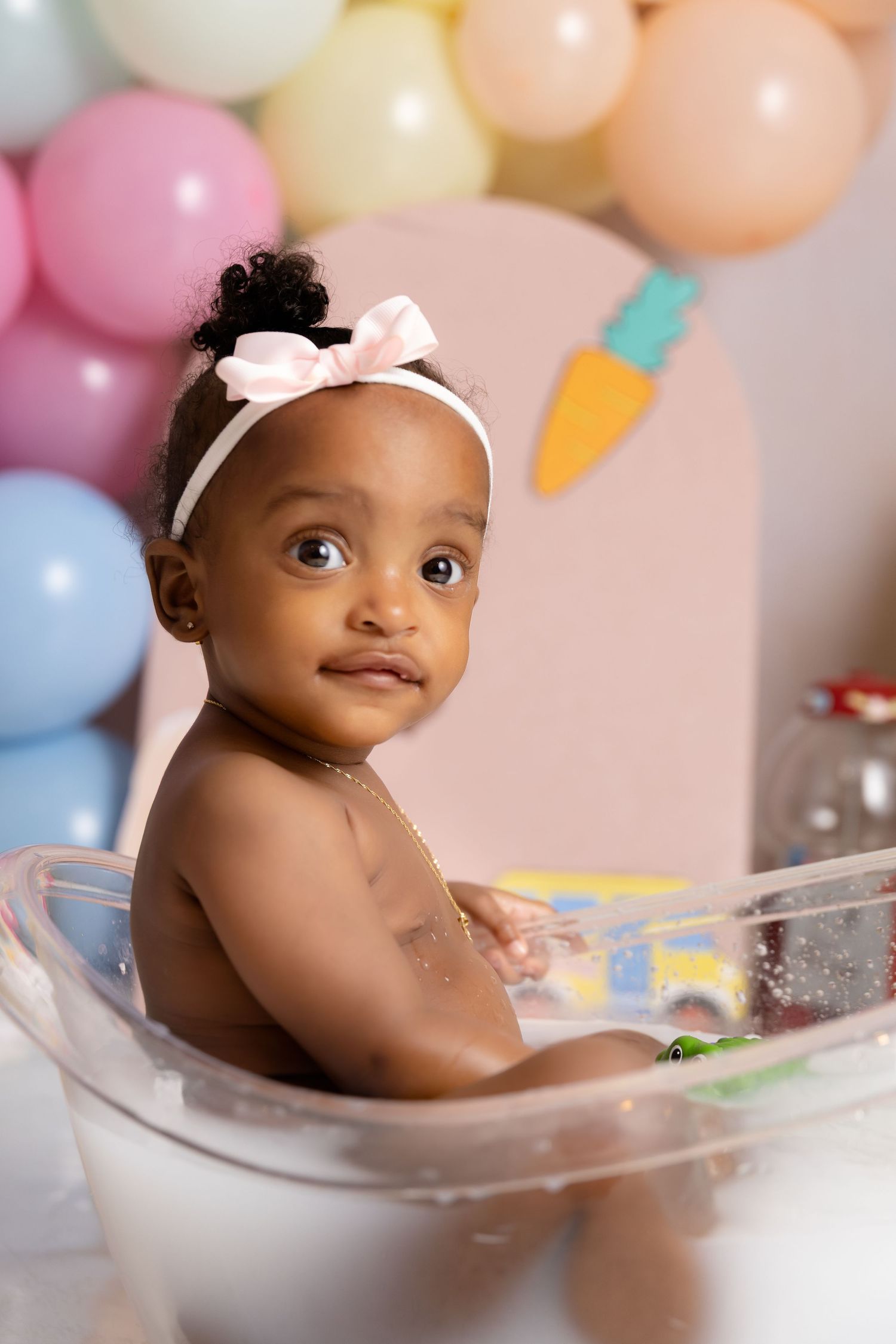 Adorable baby girl in a clear tub with a pink bow headband, surrounded by pastel balloons at a birthday celebration.