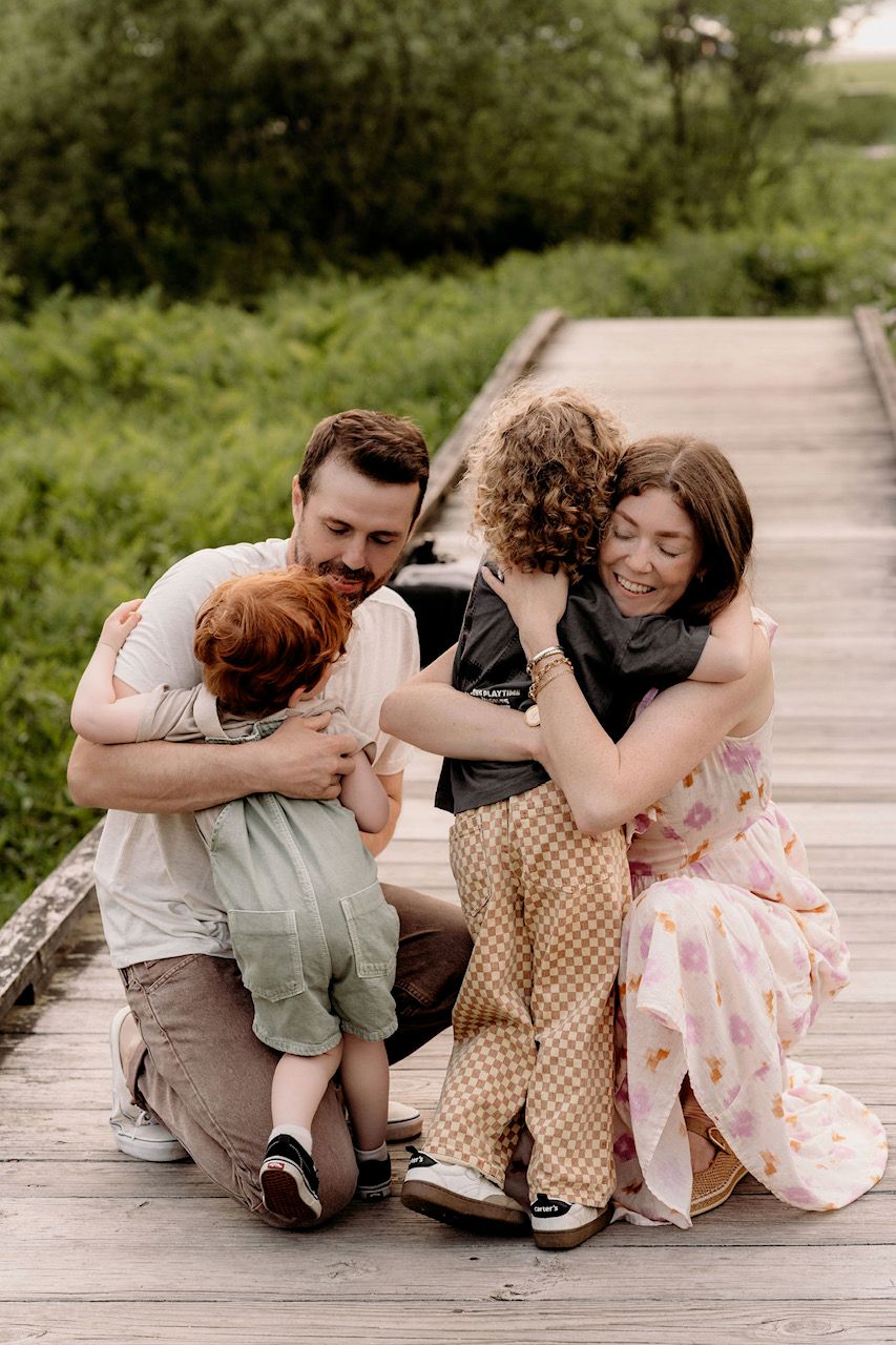 Family of four sharing warm hugs on a wooden boardwalk surrounded by lush greenery.