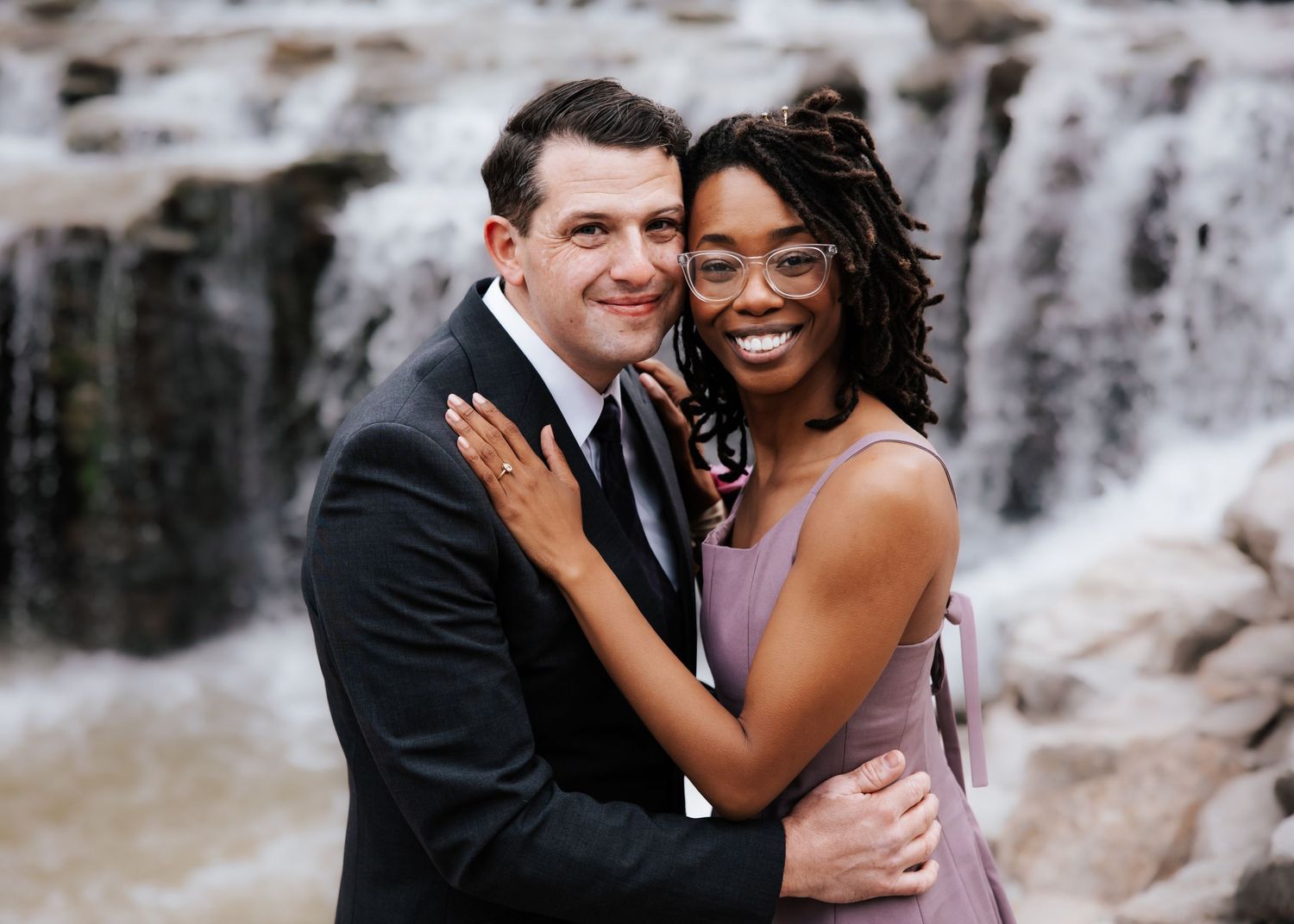 Smiling interracial couple embracing in front of waterfall, woman wearing glasses and lavender dress.